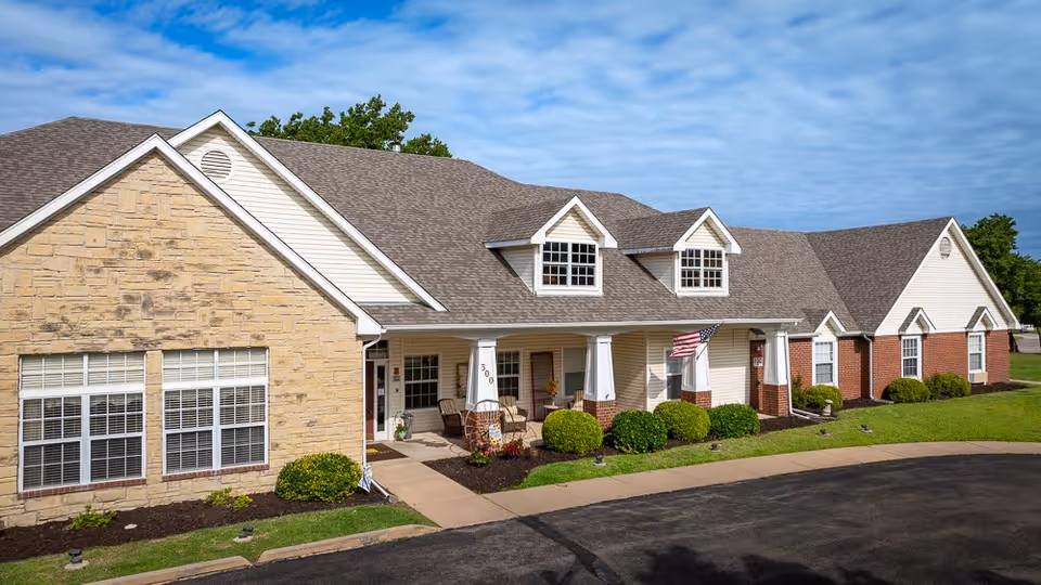 Exterior view of Homestead Assisted Living of Wellington, showing a single-story building with a combination of stone, brick, and siding facade. The building has a gray shingled roof with dormer windows, a covered porch with white columns, and an American flag displayed near the entrance. The surrounding area includes neatly trimmed bushes, a lawn, and a paved driveway.