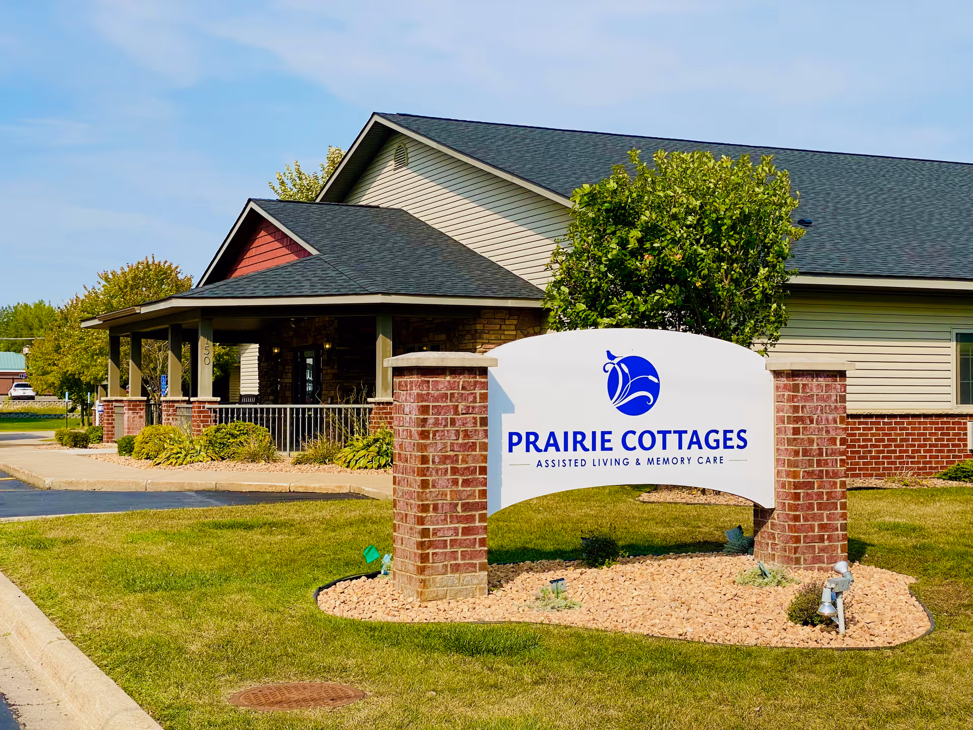 Exterior view of Prairie Cottages of Owatonna, showing a single-story building with a covered porch, brick pillars, and a sign in front that reads 'Prairie Cottages Assisted Living & Memory Care'. The area around the sign is landscaped with grass, small bushes, and rocks.