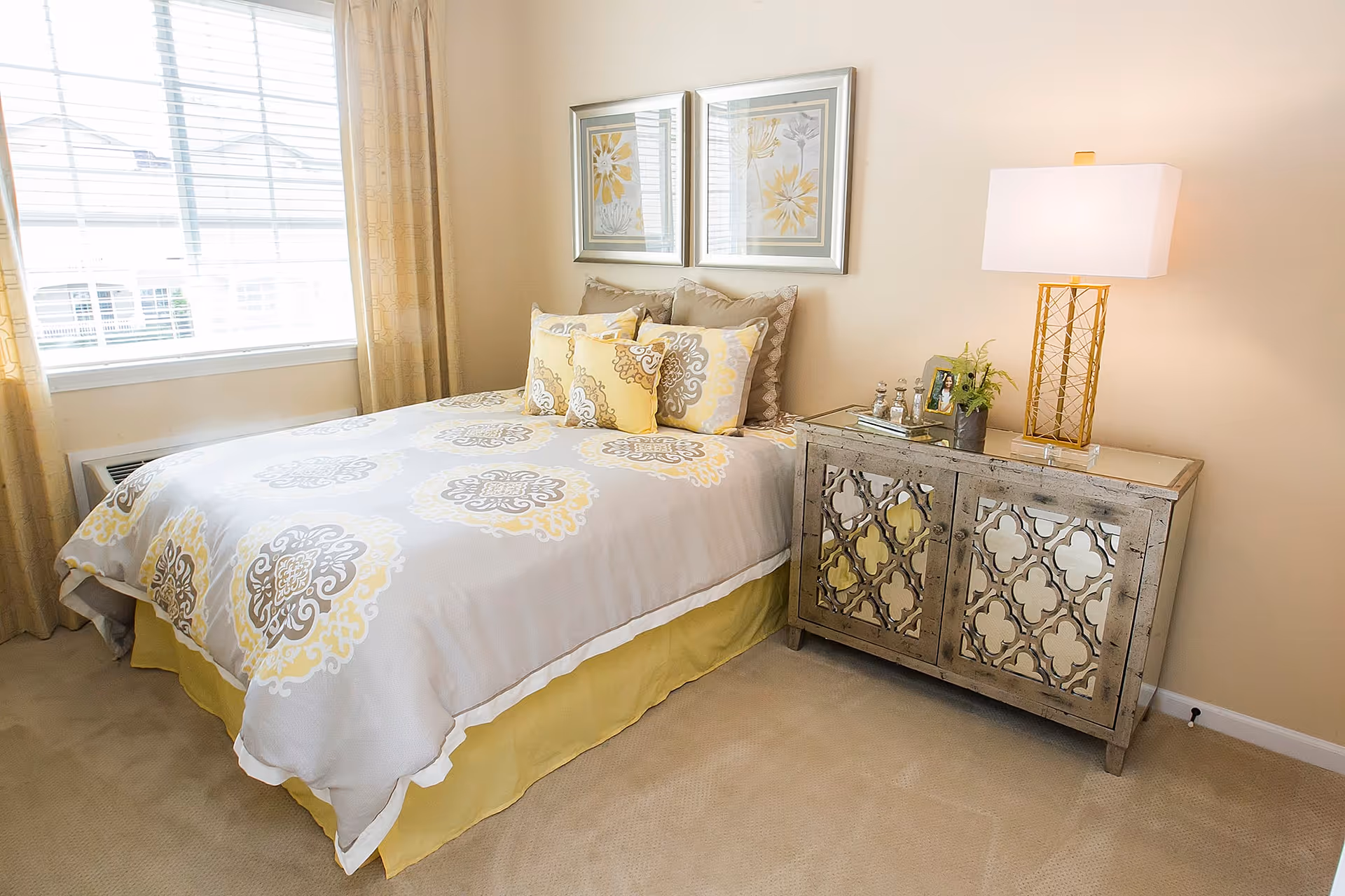 A neatly made bed with patterned bedding in shades of yellow, gray, and white, positioned next to a window with beige curtains. Beside the bed is a decorative mirrored nightstand with a gold and white table lamp, a small plant, and framed photos. Two floral-themed framed pictures hang on the wall above the bed.