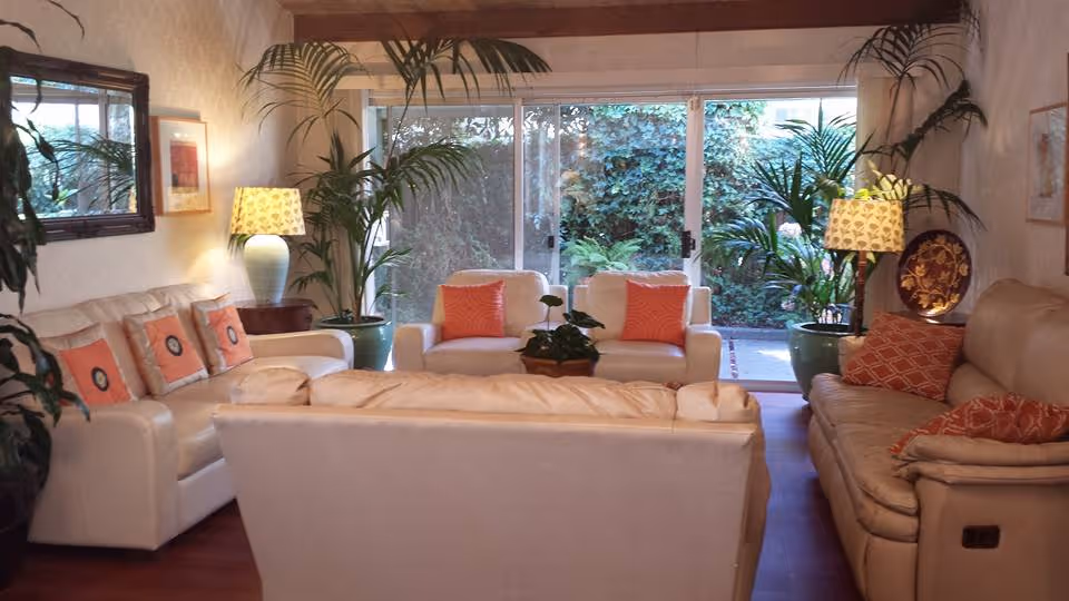 A cozy living room with three cream-colored sofas arranged around a small coffee table with a plant. The sofas have orange and patterned throw pillows. There are two table lamps with patterned shades on either side of the room, and large potted plants near sliding glass doors that lead to a garden area outside.