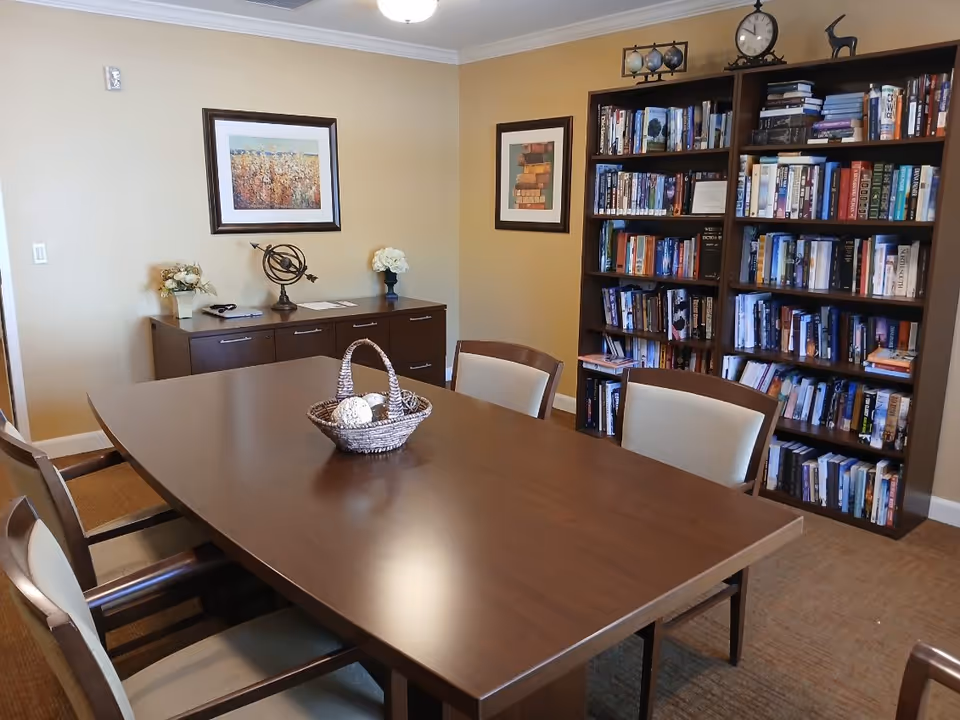 A cozy room with a large wooden table surrounded by six cushioned chairs. On the table is a woven basket with decorative balls. Against the wall is a wooden sideboard with decorative items including a globe sculpture and flower arrangements. Two framed pictures hang on the beige walls. A tall bookshelf filled with books and decorative items stands in the corner, topped with a clock and small globes.
