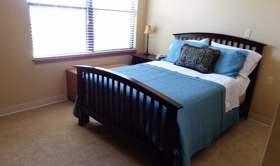 A bedroom with a wooden bed frame, neatly made with blue bedding and decorative pillows. There is a window with blinds letting in natural light, a wooden chest under the window, a nightstand with a lamp and a telephone, and beige carpeted flooring.
