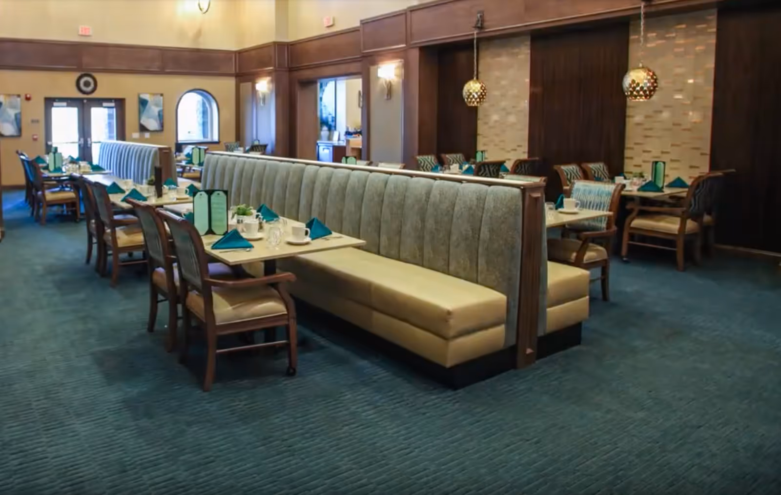 Interior view of a dining room in a senior living facility with long upholstered booth seating and wooden chairs around tables set with teal napkins, menus, cups, and glasses. The room features carpeted floors, warm lighting, and decorative wall panels.