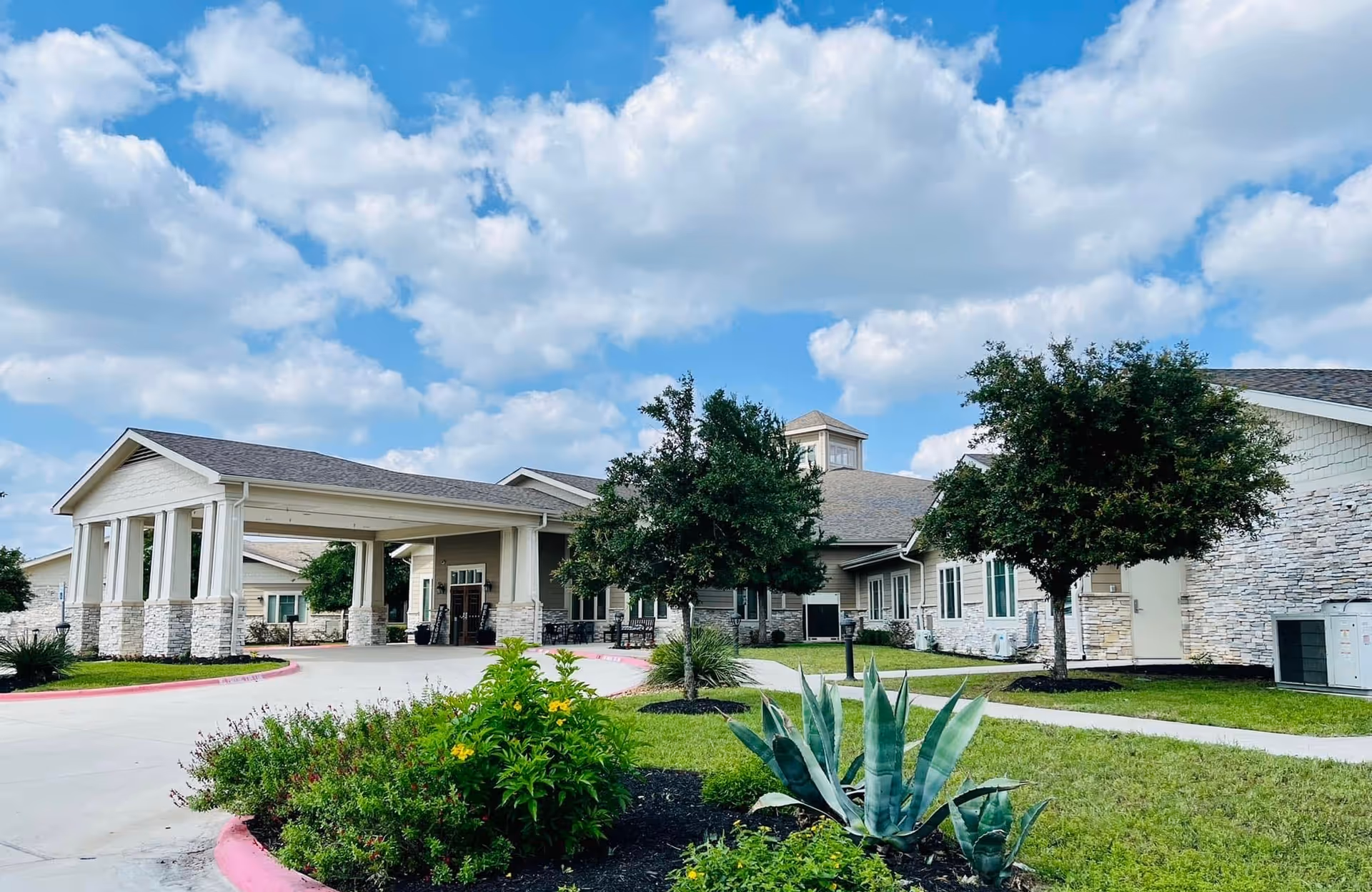Exterior view of Chapters Living of New Braunfels facility showing a single-story building with a covered entrance, surrounded by green grass, trees, and landscaped plants under a partly cloudy blue sky.