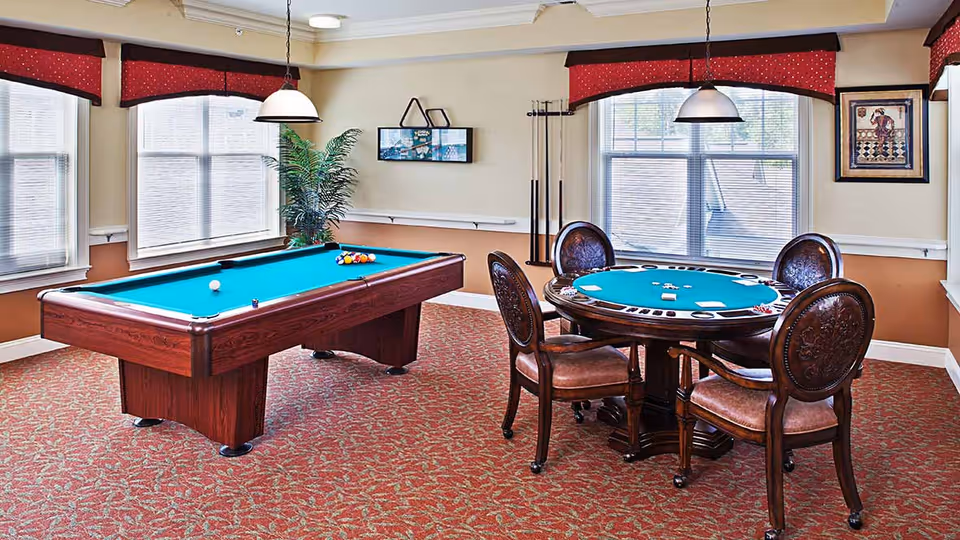 A well-lit game room featuring a pool table with balls arranged for a game and a round card table with four ornate wooden chairs. The room has large windows with blinds and red valances, a patterned carpet, a potted plant, and framed artwork on the wall.