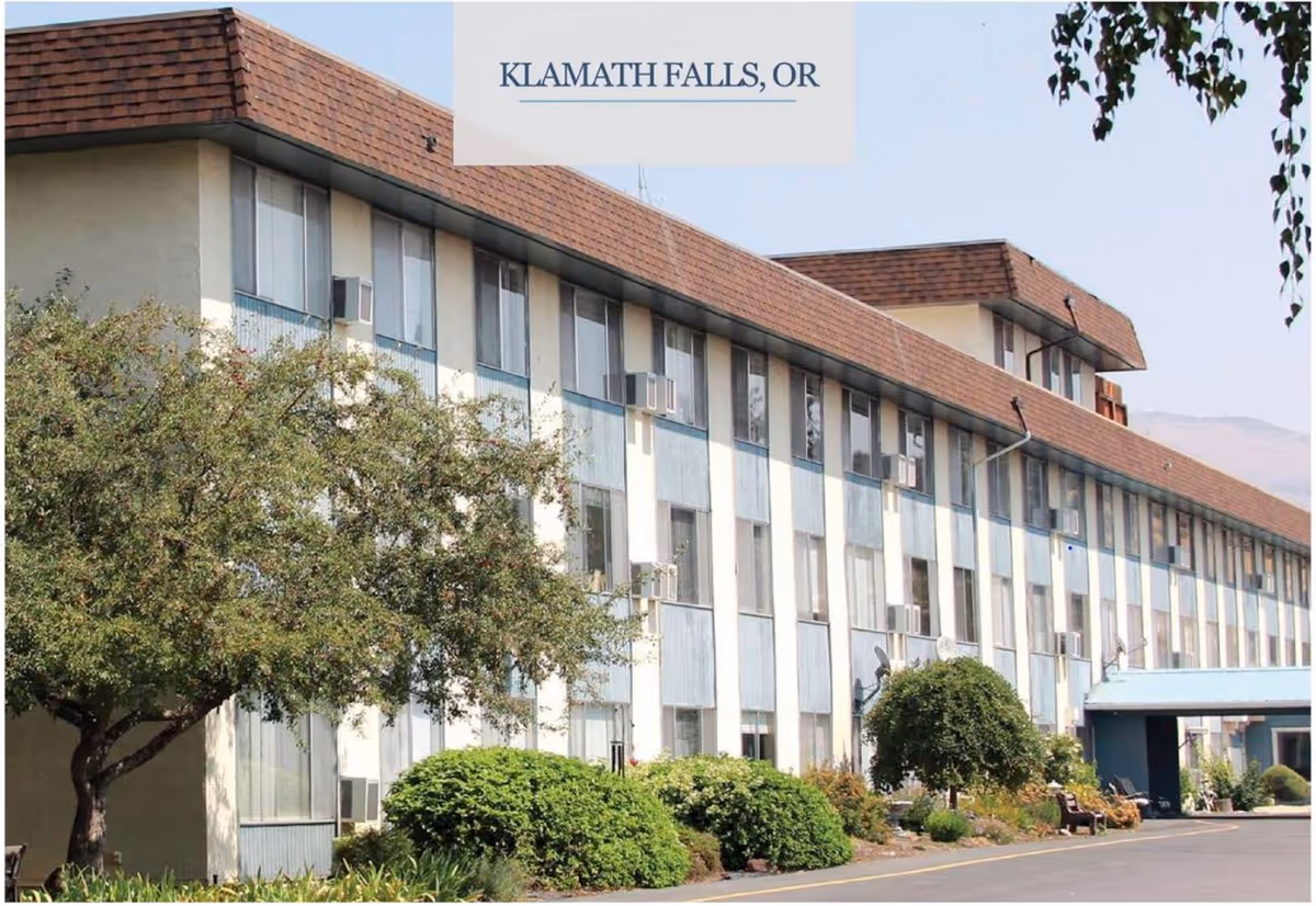 Exterior view of a multi-story senior living facility building with numerous windows and air conditioning units. The building has a brown shingled roof and light-colored walls with blue accents. There are trees and shrubs in front of the building, and a covered entrance is visible on the right side. The sky is clear.