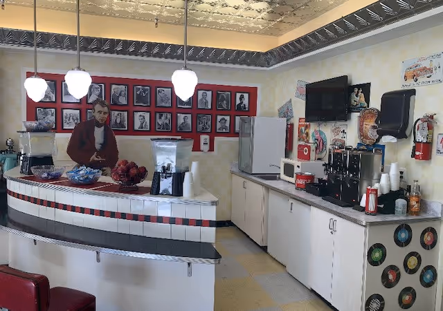Retro-style assisted-living cafe with a curved tiled service counter, pendant lights, beverage machines, refrigerator and framed wall photos.