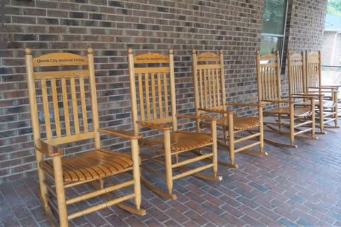 Row of wooden rocking chairs along a brick exterior wall outside Queen City Assisted Living
