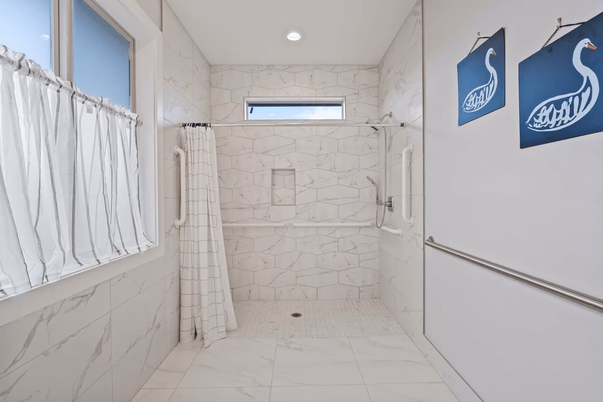 A spacious bathroom with a walk-in shower featuring white and light gray marble-patterned tiles. The shower has grab bars on the walls and a handheld showerhead. There is a window with white curtains on the left wall and two blue wall hangings with white swan designs on the right wall. A metal handrail runs along the right side wall.