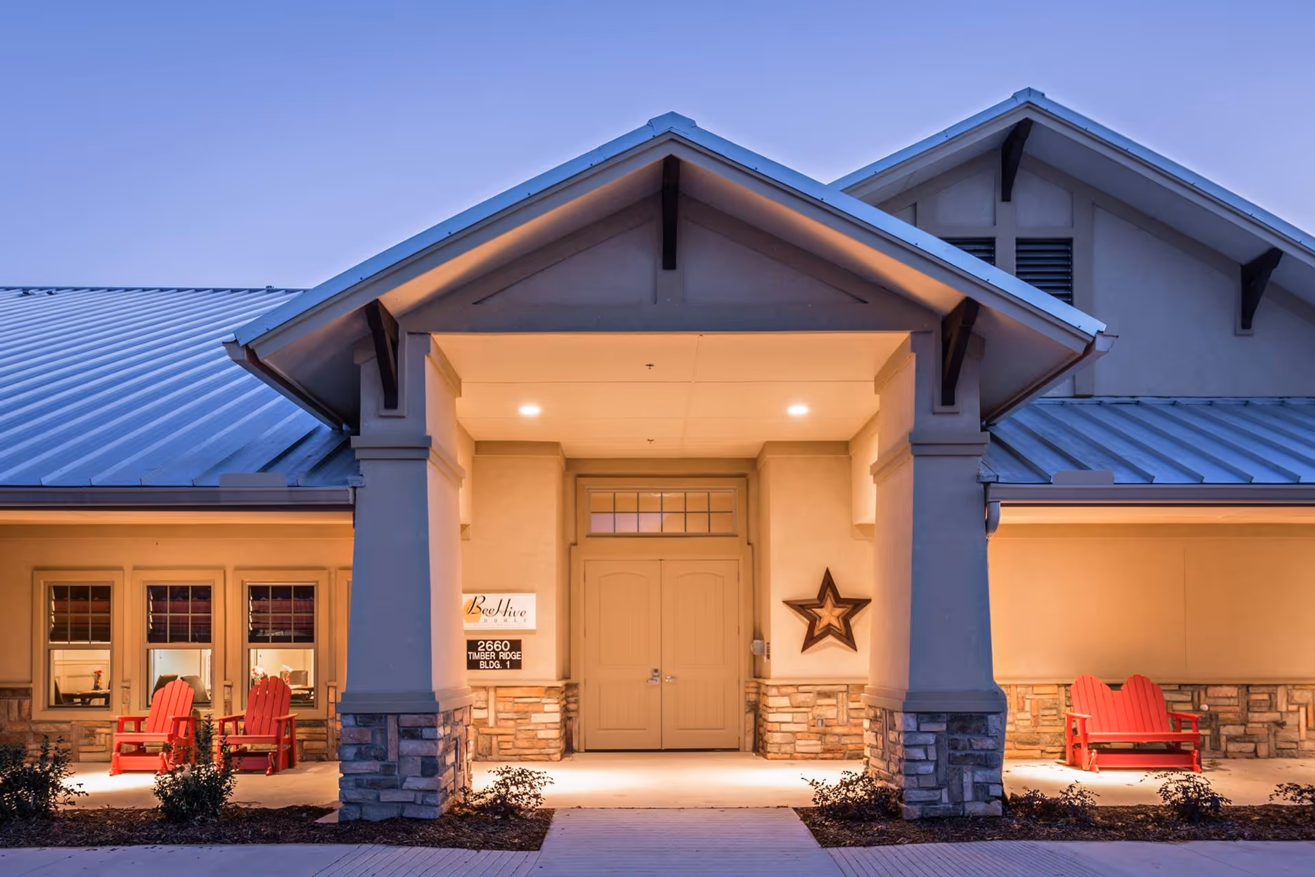 Exterior front entrance of BeeHive Homes of Frisco at dusk, featuring a covered porch with stone pillars, double doors, a decorative star on the wall, and red Adirondack chairs on either side.