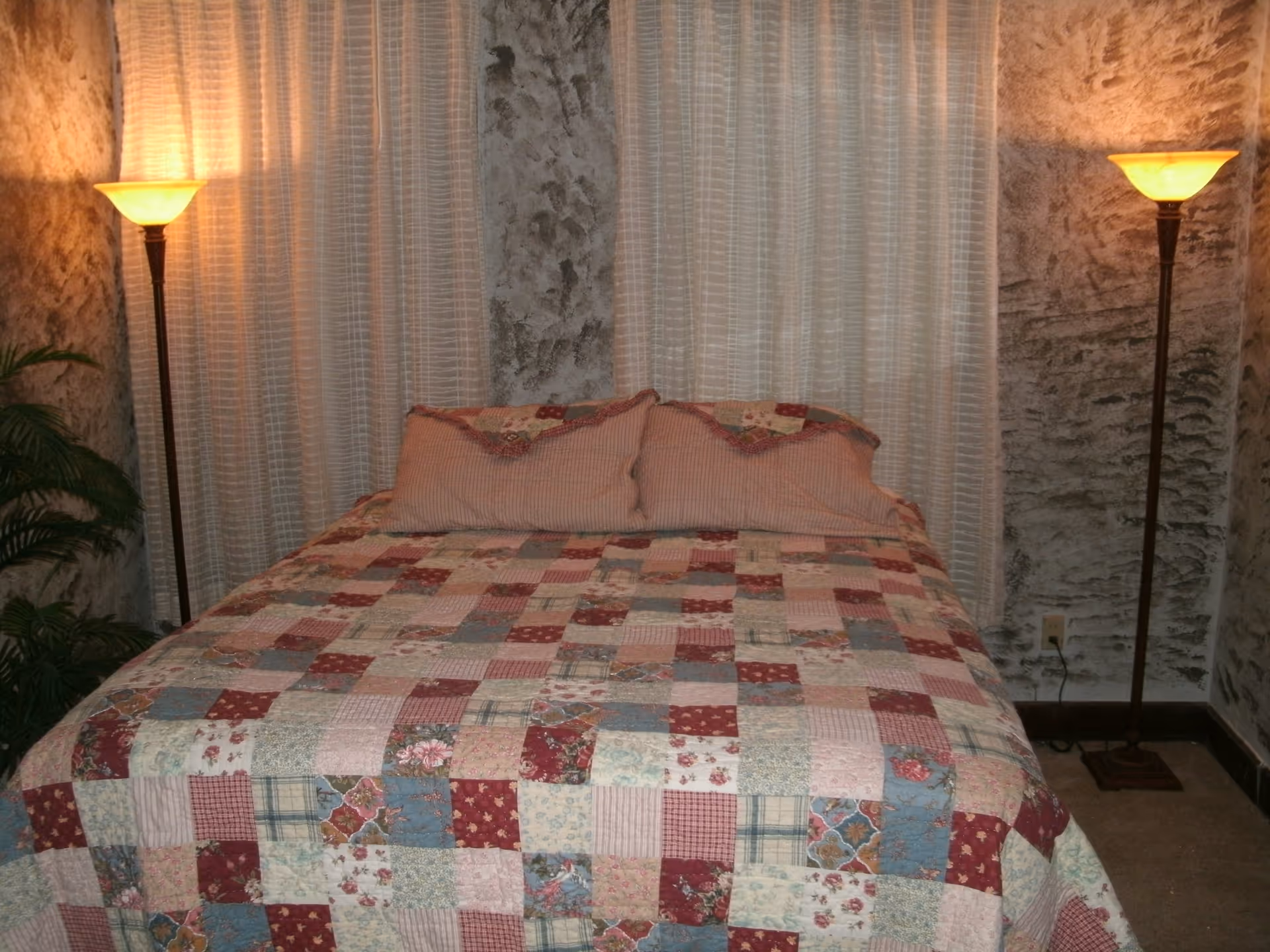 A bedroom with a bed covered in a patchwork quilt featuring various patterns and colors including red, blue, and beige. Two matching pillows rest against the headboard. The bed is flanked by two tall floor lamps with warm yellow light. The walls have a textured gray finish and there are sheer white curtains behind the bed. A green plant is partially visible on the left side.
