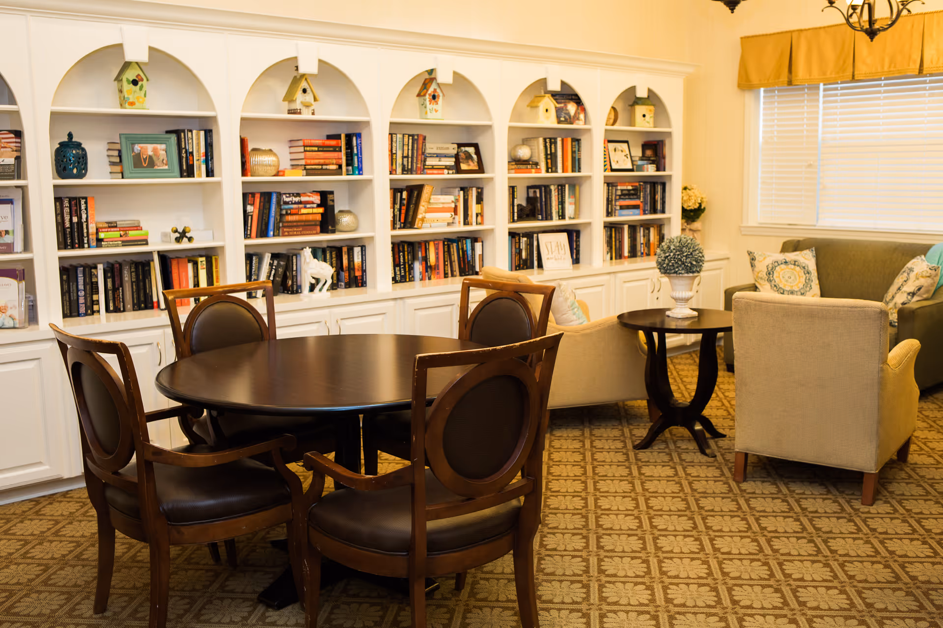 Cozy common room featuring a round wooden table with chairs, armchairs and a sofa arranged before built-in bookshelves.