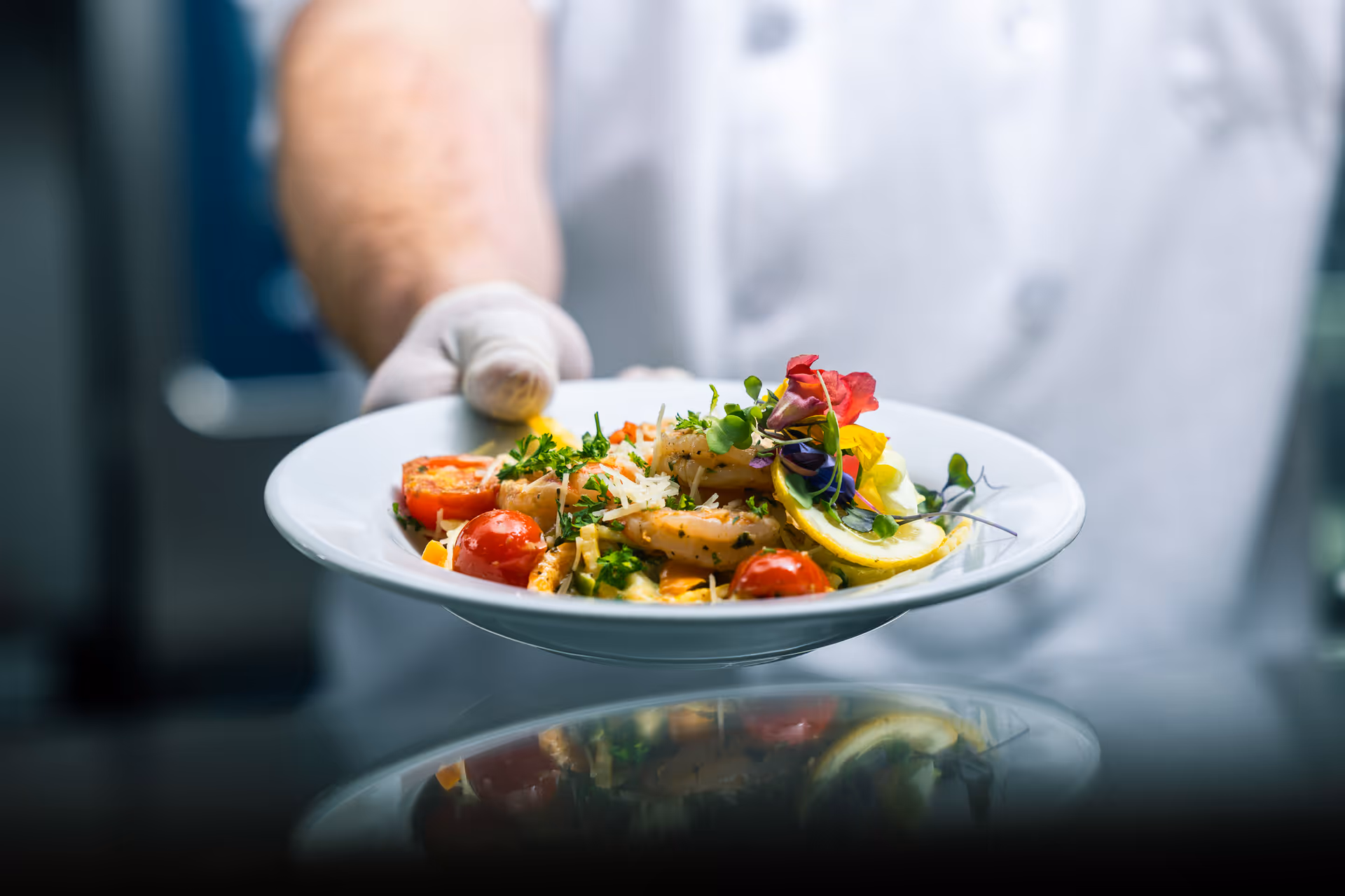 A chef wearing a white uniform and glove is holding out a white plate with a gourmet dish consisting of shrimp, cherry tomatoes, lemon slice, herbs, and edible flowers.