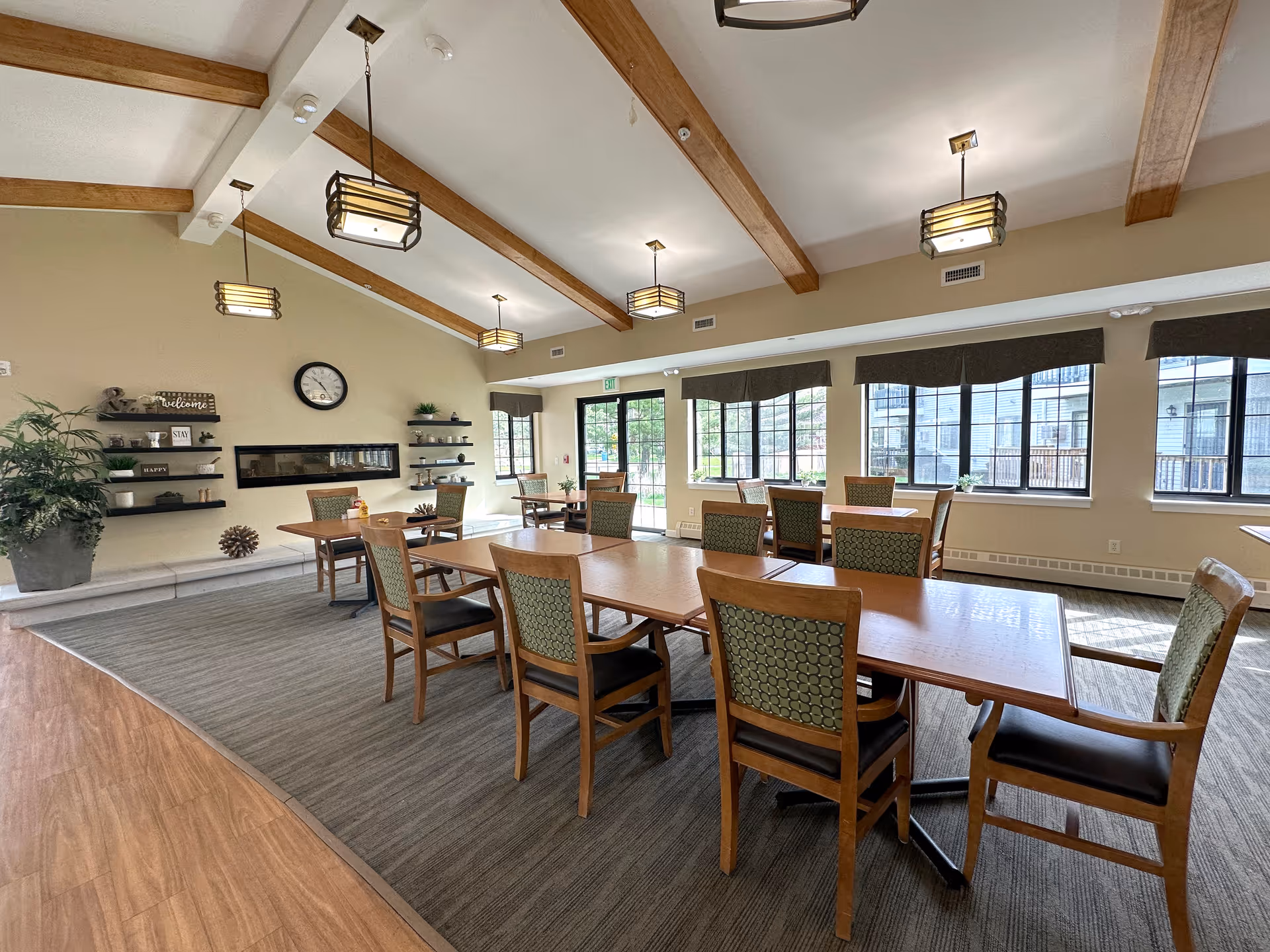 A bright and spacious dining room with wooden tables and chairs arranged neatly on a carpeted floor. The room features large windows with dark valances allowing natural light to fill the space. The ceiling has exposed wooden beams and modern hanging light fixtures. On one wall, there are decorative shelves with plants and signs, and a clock above a built-in fireplace.
