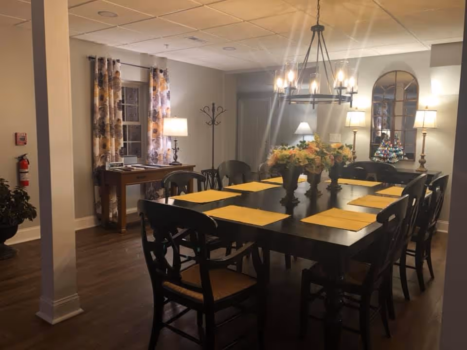 Interior dining room with a long dark wood table set with yellow placemats, floral centerpieces, chandelier, and lamps.