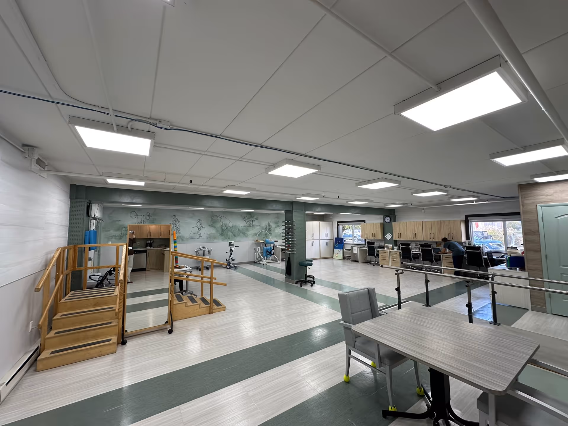 Open rehabilitation gym interior with parallel bars, practice stairs, therapy equipment, desks and tables.