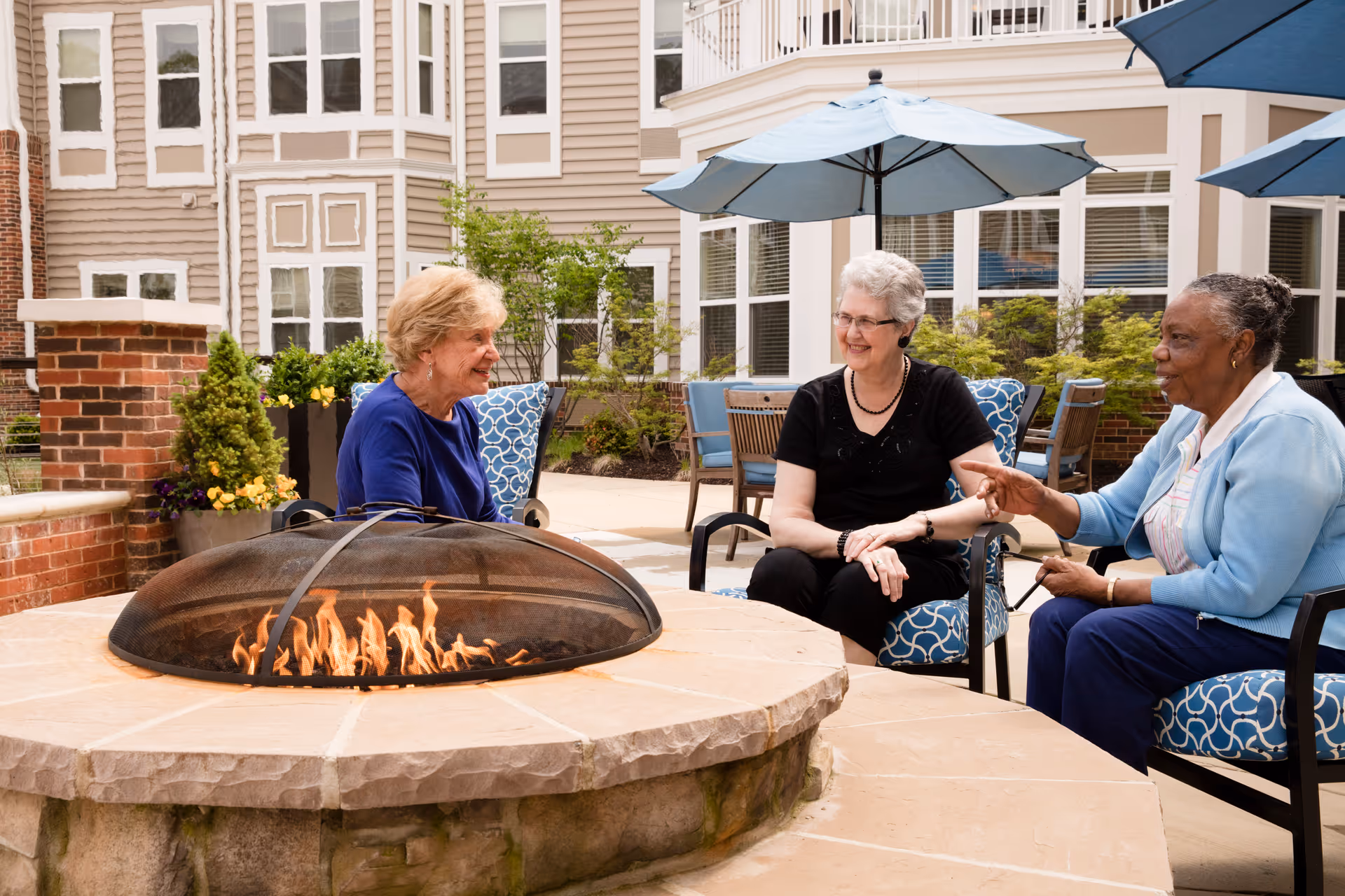 Three elderly women sitting around a stone fire pit with a protective mesh cover in an outdoor patio area of a senior living facility. They are engaged in conversation, seated on cushioned chairs with blue and white patterned fabric. The background shows a multi-story building with large windows, potted plants, and blue patio umbrellas providing shade.