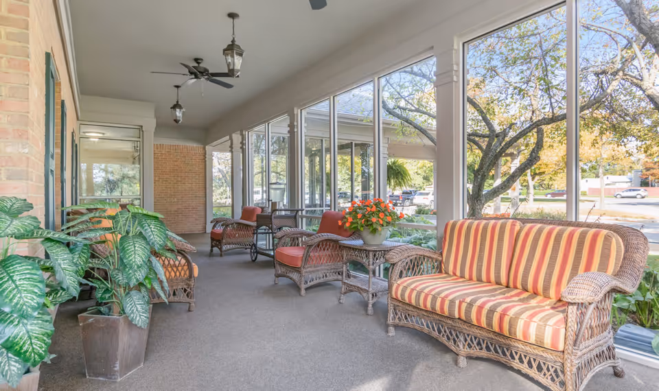 A bright and airy enclosed porch area with wicker furniture including a striped cushioned sofa, armchairs with orange cushions, and a small table with a potted flowering plant. Large windows line one side, offering a view of trees and parked cars outside. Ceiling fans and hanging lantern-style lights are mounted on the ceiling. Several potted green plants are placed along the brick wall on the left.