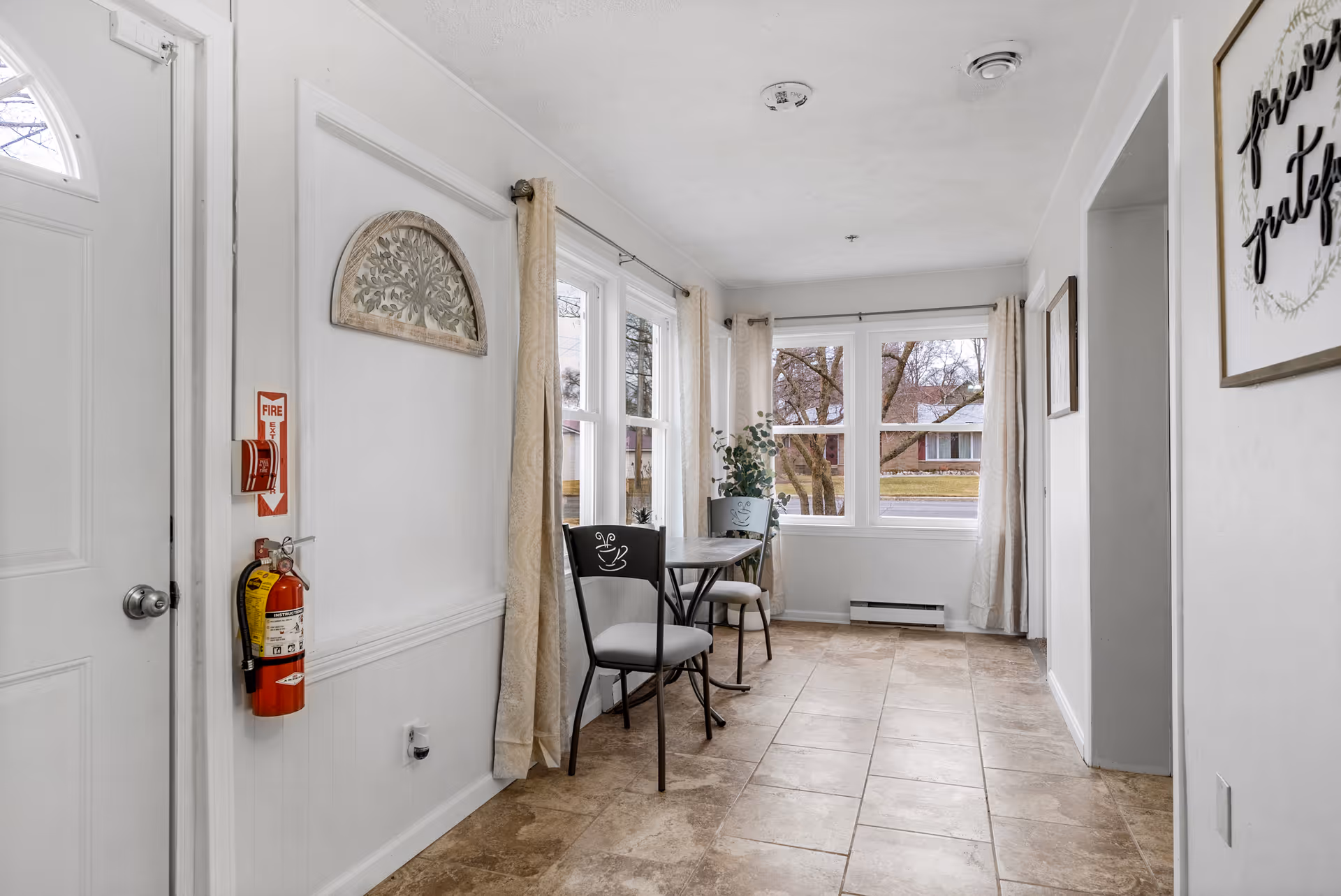 A bright and clean interior hallway with large windows letting in natural light. There is a small table with two chairs near the windows, beige curtains, a fire extinguisher mounted on the wall, and a framed sign that reads 'forever grateful'. The floor is tiled and the walls are painted white.