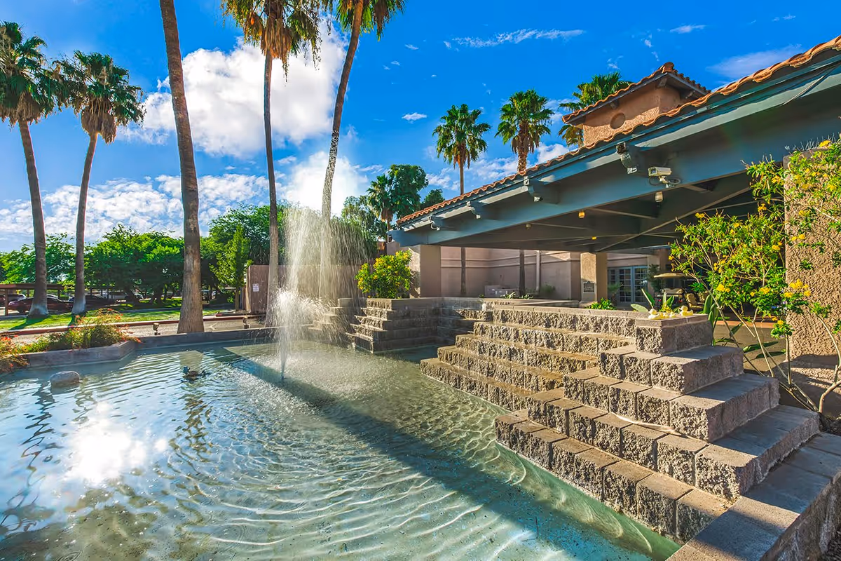 Outdoor view of The Fountains at La Cholla facility featuring a water fountain in a shallow pool with stone steps, surrounded by palm trees and greenery under a partly cloudy blue sky.