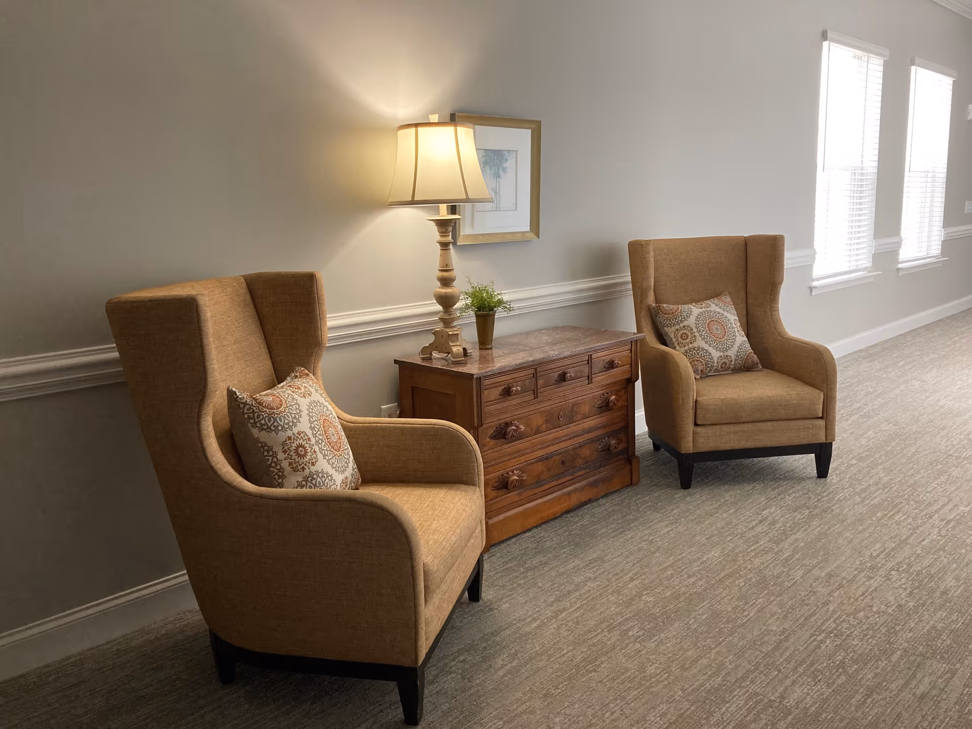 Two brown upholstered armchairs with patterned cushions are placed on either side of a wooden chest of drawers with a table lamp and a small potted plant on top. The setting is a well-lit hallway with three windows on the right side, beige walls, and carpeted floor.