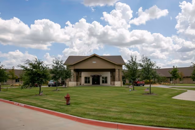 Front exterior of a one-story brick building with a covered main entrance, landscaped lawn, and driveway beneath a partly cloudy sky.