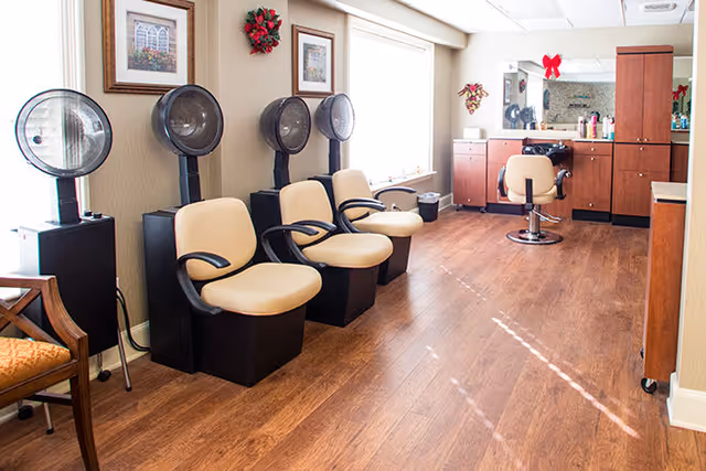 Interior view of a salon area with four beige salon chairs, each paired with a hair dryer hood. The room has wooden flooring, light-colored walls, framed artwork, and a large window letting in natural light. There is a counter with a mirror and salon supplies in the background, decorated with a red bow and holiday wreaths.