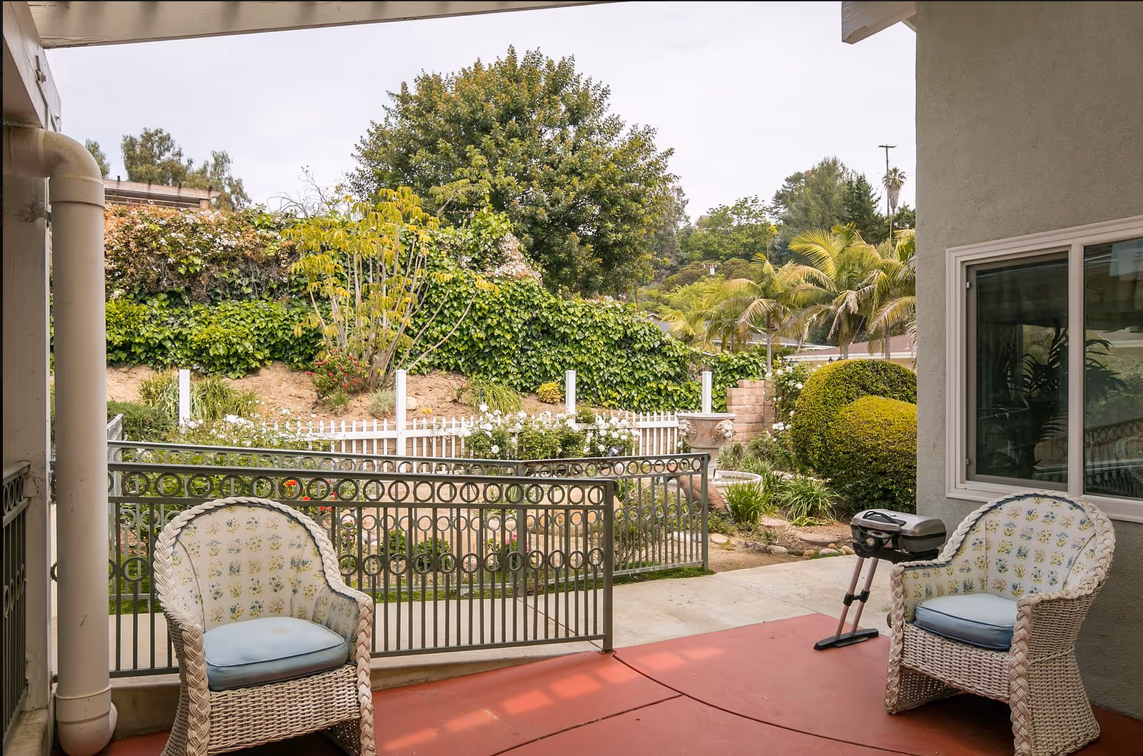 Covered outdoor patio area with two wicker chairs featuring floral cushions, a small grill, and a view of a garden with various green plants, bushes, and trees surrounded by a metal railing and a white picket fence.