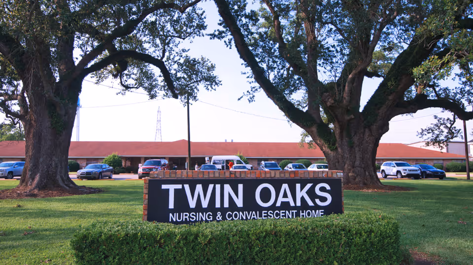 Exterior view of Twin Oaks Nursing & Convalescent Home with a large sign in front on a grassy area, flanked by two large oak trees and several parked cars in the background.