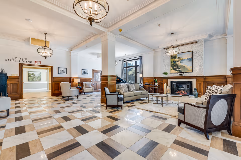 Spacious and elegant senior living facility common area with checkered tile floor, comfortable seating including sofas and armchairs, a fireplace with a painting above it, large windows with curtains, and decorative ceiling lights.