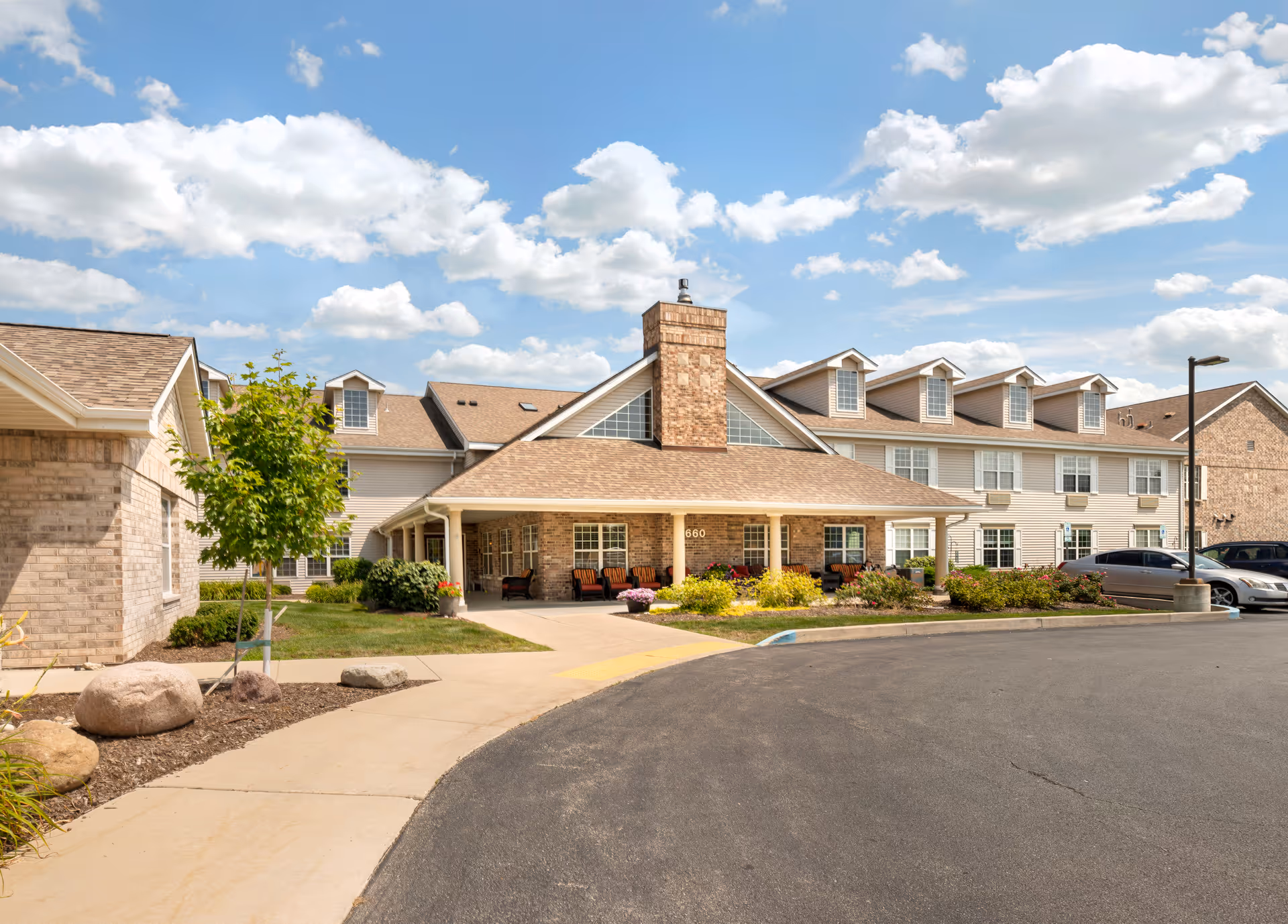 Front entrance of a two-story senior living building with a covered porte-cochère, landscaped flower beds, and parked cars under a blue sky.