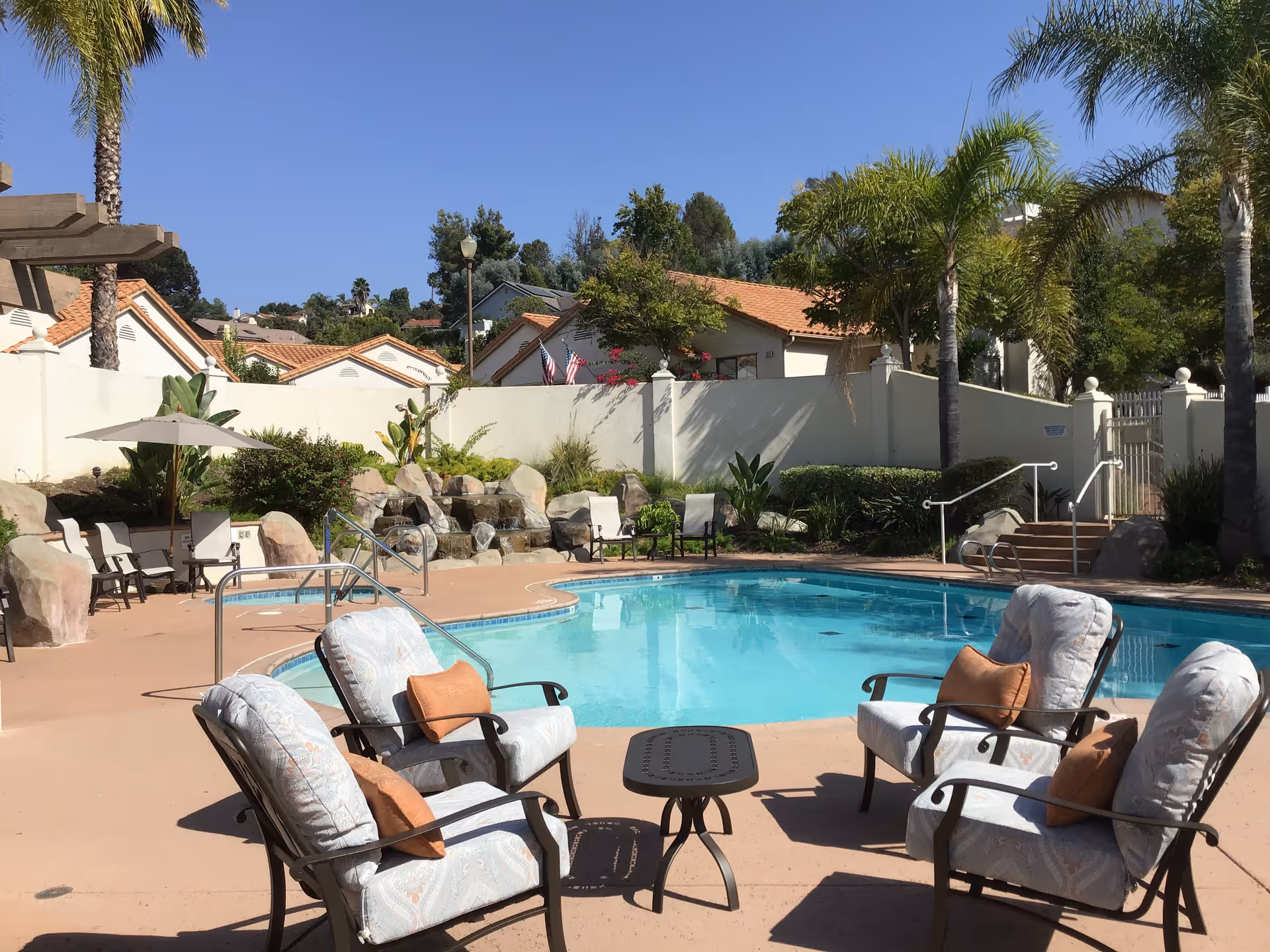 Outdoor swimming pool area with four cushioned chairs and a small table arranged around the pool. Palm trees and other greenery surround the pool, with a waterfall feature and rock landscaping in the background. Residential-style buildings with tiled roofs are visible behind a white wall under a clear blue sky.