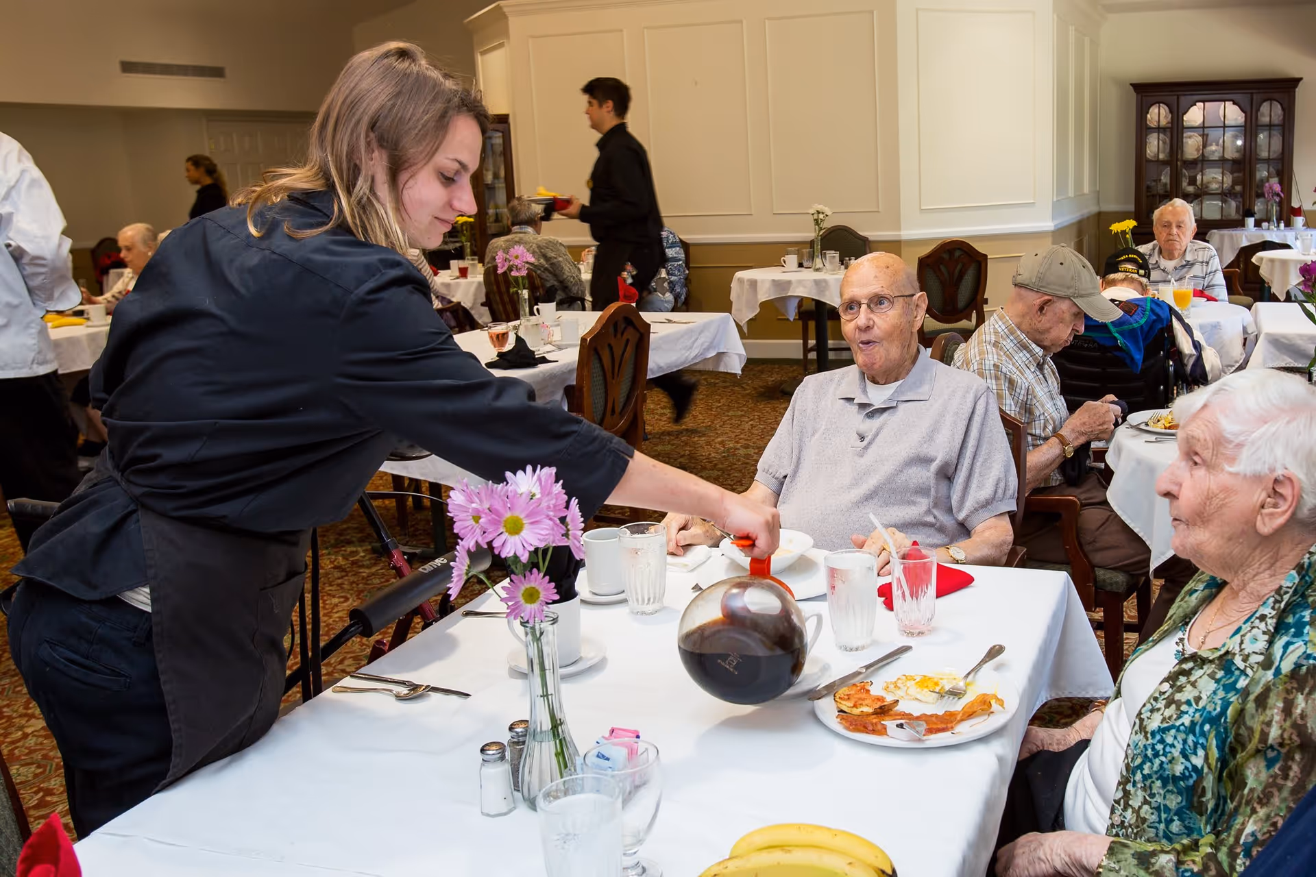 A young female server pours coffee for an elderly man seated at a dining table in a senior living facility dining room. Other elderly residents are seated at nearby tables, eating and conversing. The room has white walls, wooden chairs, and tables covered with white tablecloths and decorated with flowers.