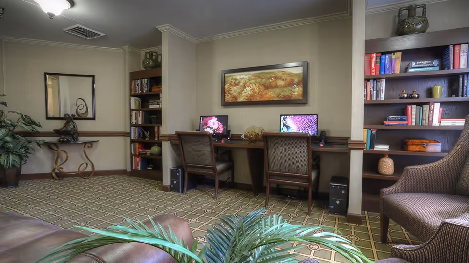 A cozy senior community room featuring two desktop computers on a wooden desk with two chairs. The room has bookshelves filled with books and decorative items on either side of the desk. There is a framed landscape painting on the wall above the desk, a small table with a sculpture and a mirror on the left side, and comfortable seating with plants in the foreground. The carpet has a geometric pattern and the walls are painted beige with wooden trim.