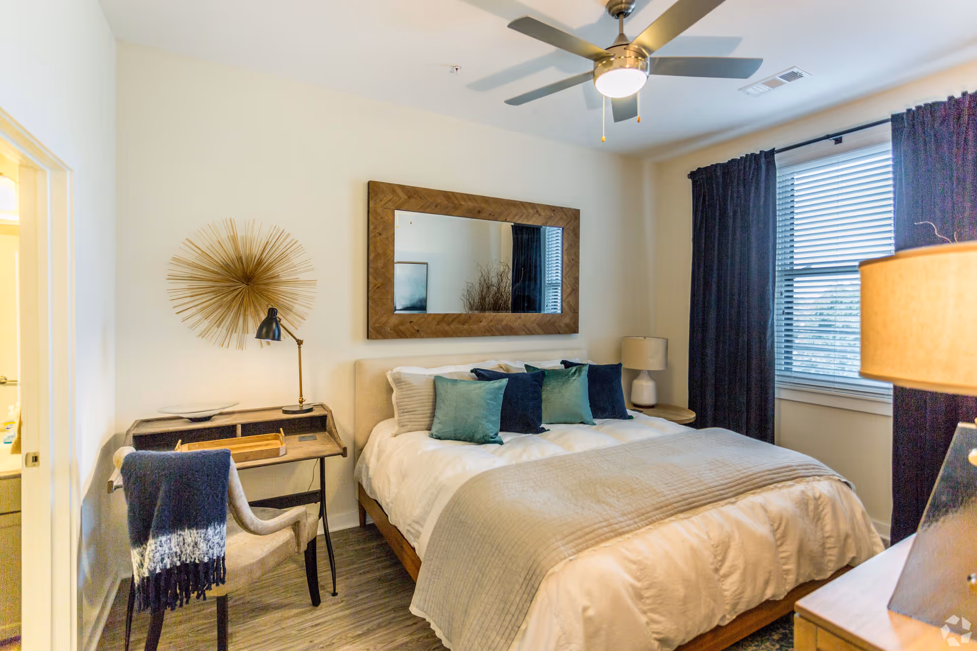 A cozy bedroom with a large bed featuring white bedding and multiple blue and green pillows. Above the bed is a large wooden framed mirror. To the left of the bed is a small wooden desk with a black desk lamp and a beige chair draped with a blue and white throw. A decorative sunburst wall art is mounted above the desk. On the right side of the bed is a round wooden nightstand with a white lamp. The room has light-colored walls, a ceiling fan with a light, and a window with dark curtains and blinds.
