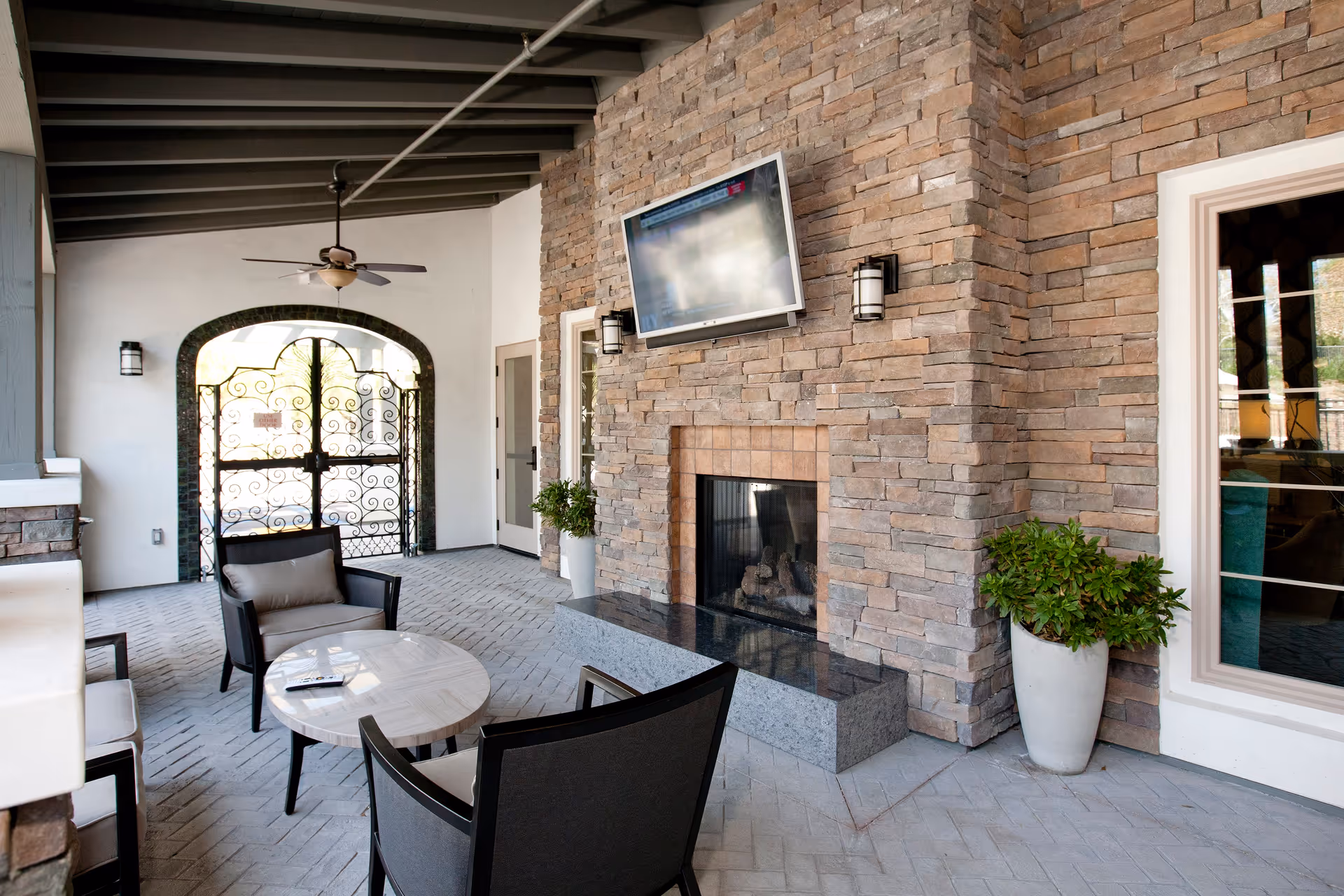 Covered outdoor seating area with a stone fireplace, a mounted flat-screen TV above the fireplace, two cushioned chairs around a round table with a remote control on it, a ceiling fan, potted plants, and a decorative wrought iron gate in the background.