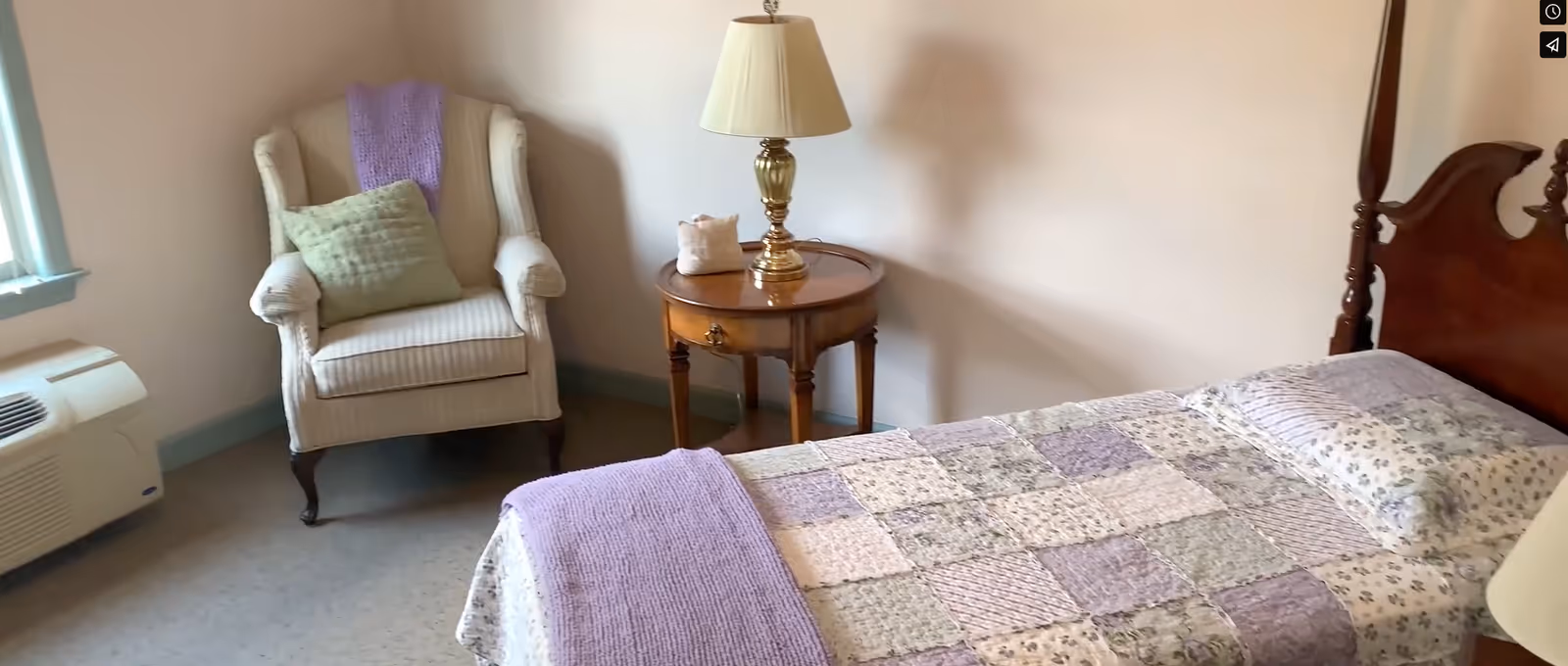 A cozy bedroom featuring a wooden four-poster bed with a patchwork quilt in shades of purple and cream. Next to the bed is a round wooden side table with a brass lamp and a small decorative item. An upholstered armchair with a green pillow and a purple throw blanket is positioned near a window with light blue trim. A white air conditioning unit is visible below the window.