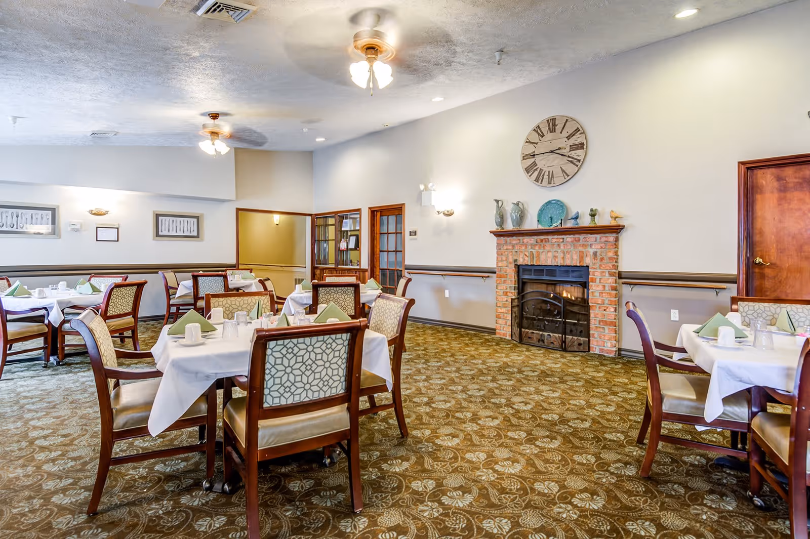 Dining room with multiple tables covered with white tablecloths, set with cups, glasses, and green folded napkins. The room features a patterned carpet, a brick fireplace with decorative items on the mantel, a large wall clock above the fireplace, ceiling fans with lights, and framed artwork on the walls.