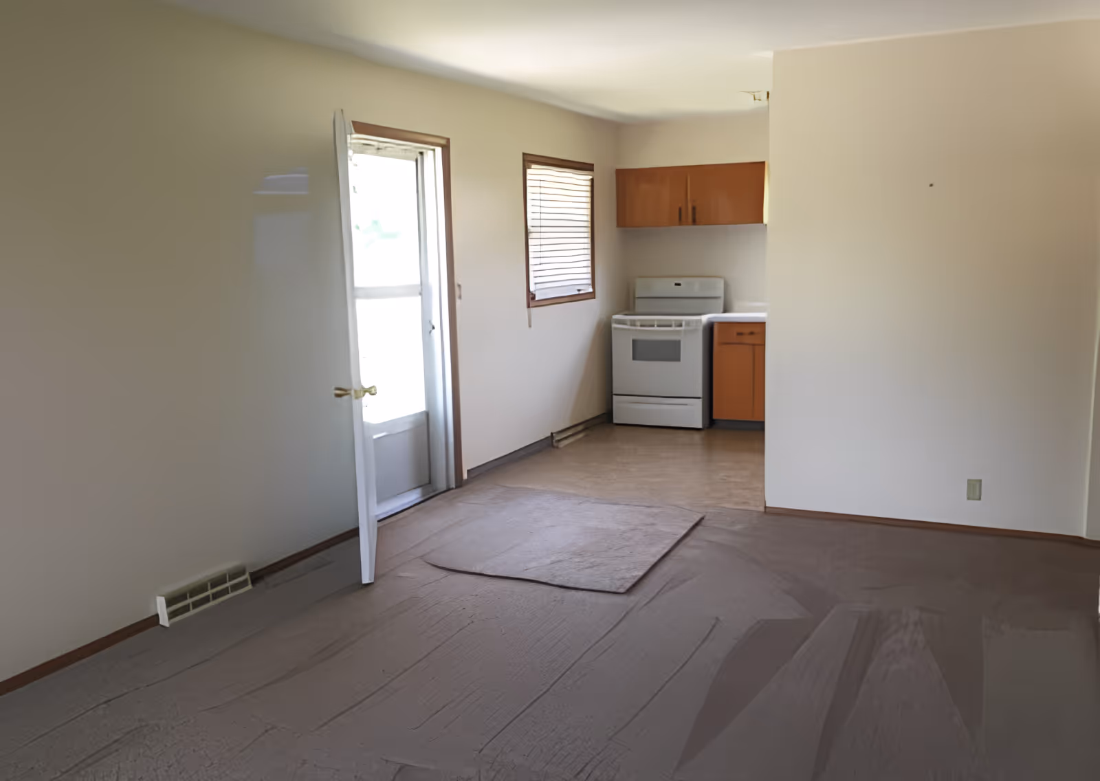 An empty interior room with beige walls and carpeted floor, featuring a small kitchen area with a white stove, wooden cabinets, and a window with blinds. There is a door with a window panel that is partially open, allowing natural light into the room.