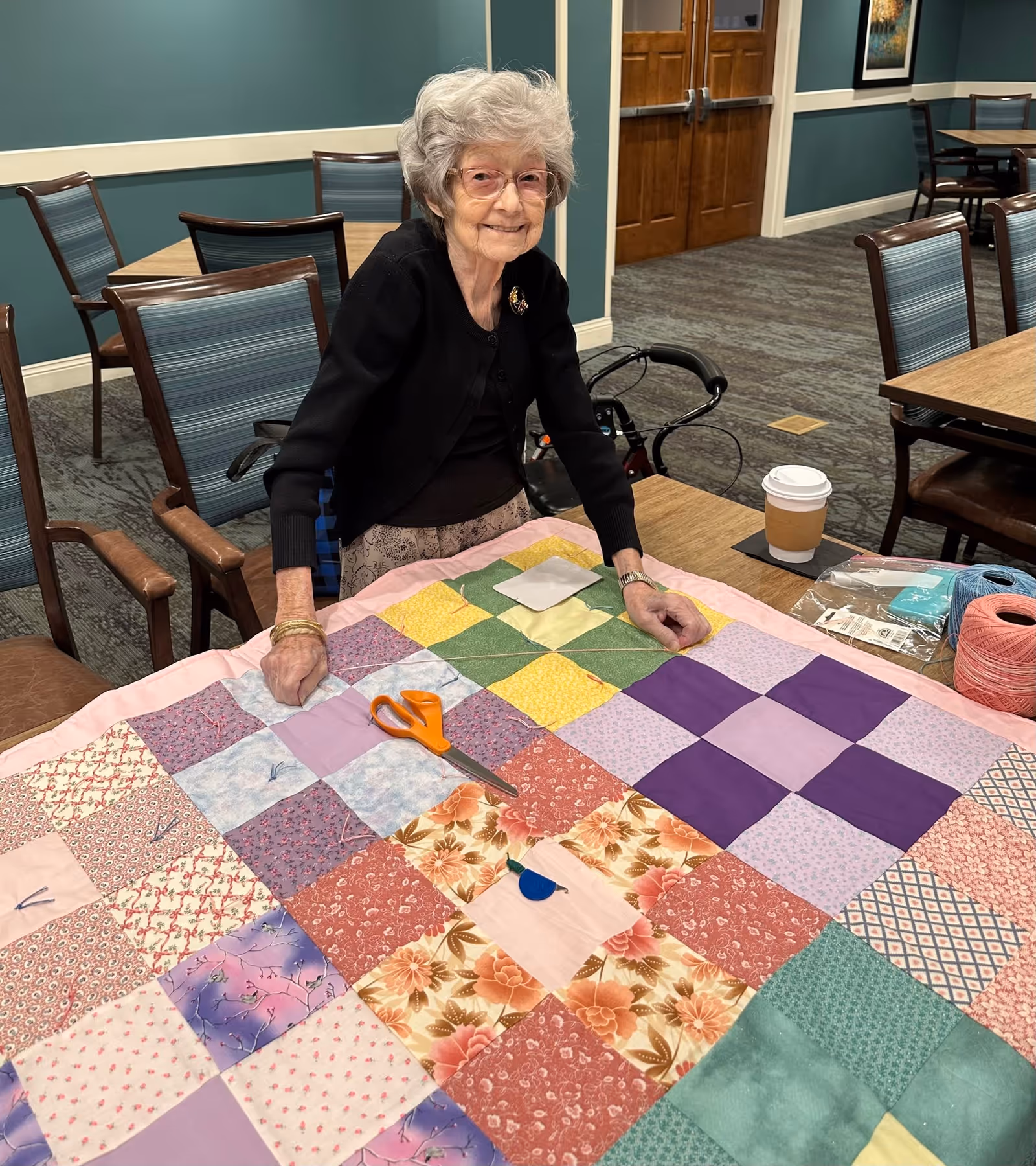 An older woman smiling as she works on a colorful patchwork quilt at a table in a senior living community dining/activity room.