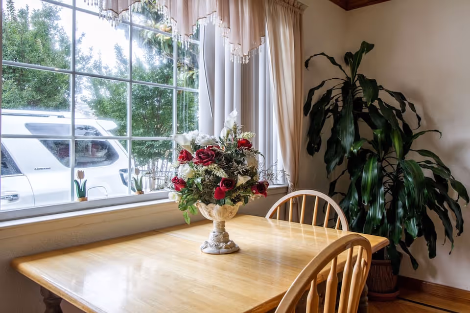 A wooden dining table with a floral centerpiece featuring red and white flowers is placed near a large window with white curtains. Two wooden chairs are positioned around the table, and a large green potted plant is in the corner of the room. Outside the window, a white vehicle and green trees are visible.