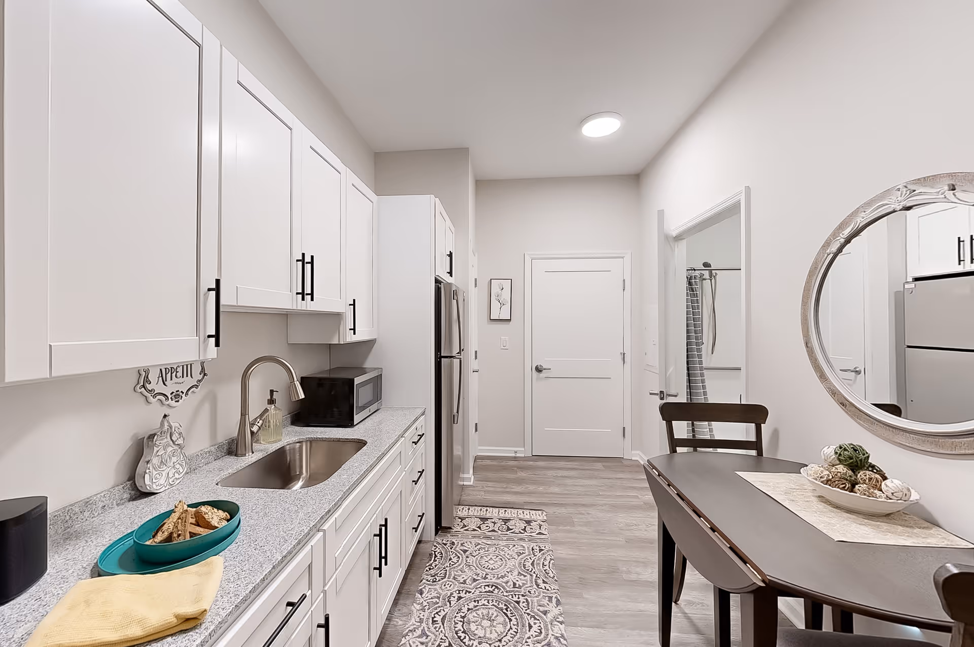A modern kitchen with white cabinets, a granite countertop, a stainless steel sink, and a microwave. There is a small dining table with two chairs and a decorative bowl with woven balls on a placemat. A round mirror hangs on the wall above the table. The floor has a patterned rug and light wood flooring. A door is visible at the end of the kitchen, and a bathroom with a shower curtain is partially visible through an open door.