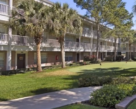 Three-story apartment building with balconies, palm trees, and a grassy courtyard with a walkway.