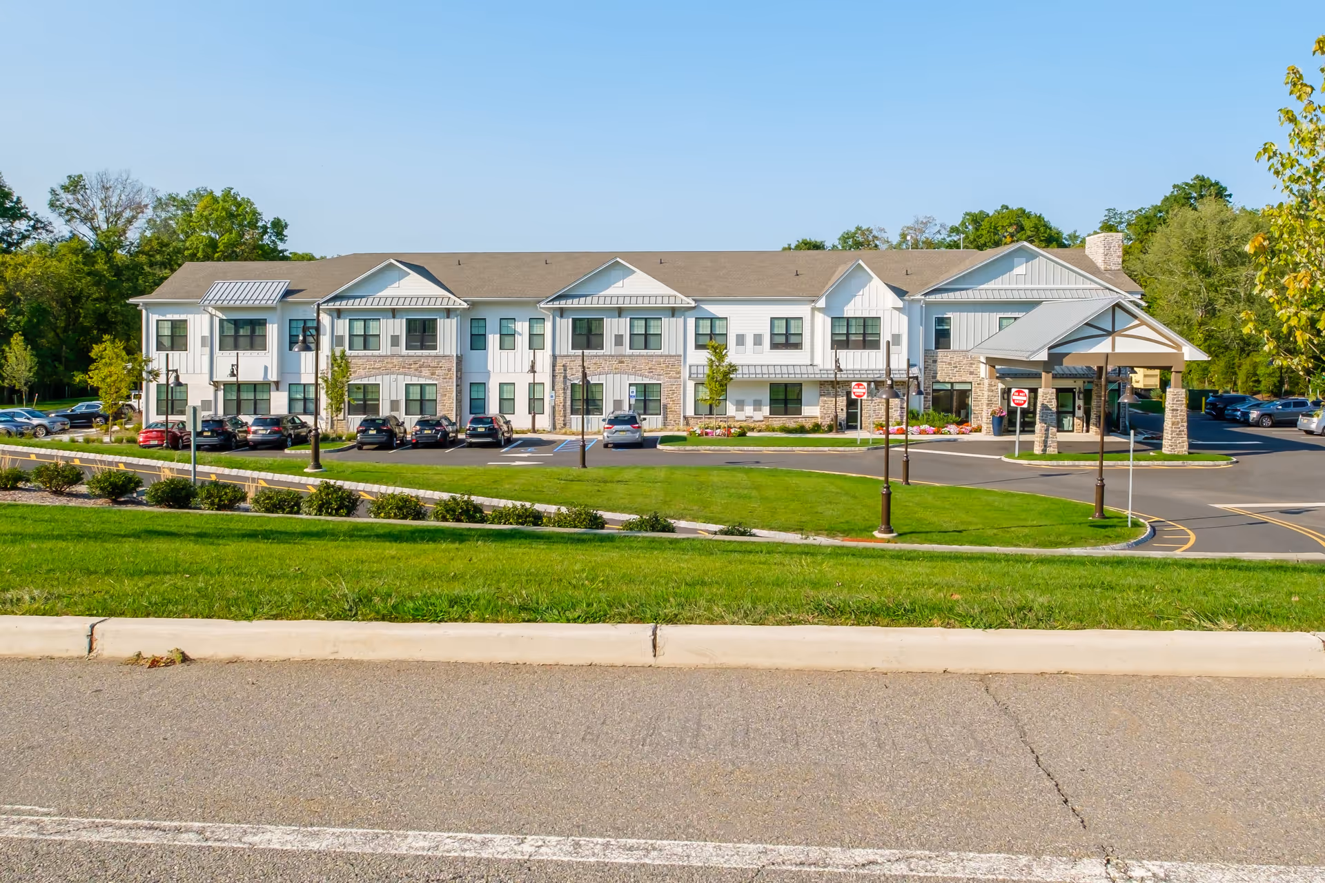 Front view of a two-story senior living building with a covered entrance, parking lot, and landscaped lawn.