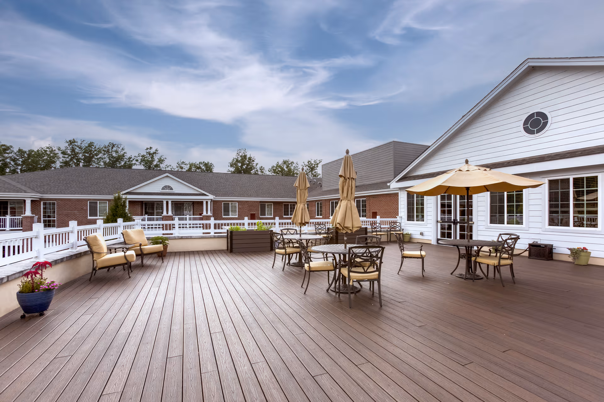 Outdoor patio area with wooden flooring, several tables and chairs with beige cushions, and large beige umbrellas. The patio is adjacent to a white building with large windows and a circular window near the roof. In the background, there is a brick building with white railings and trees under a partly cloudy sky.