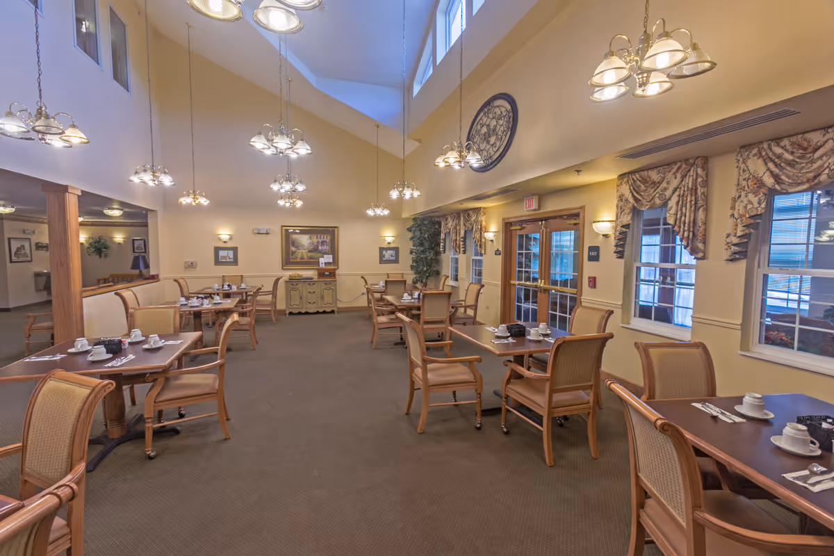 Bright dining room with multiple wooden tables and chairs set for service under a vaulted ceiling with hanging chandeliers.