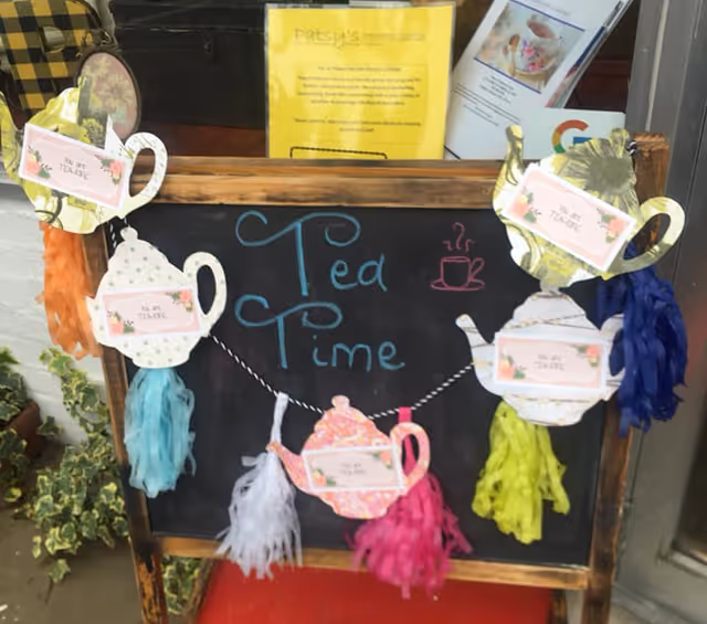A small chalkboard sign with the words 'Tea Time' written in blue and a pink steaming cup drawn on it. The board is decorated with paper cutouts of teapots and teacups, each with a label that says 'It's Tea Time,' and colorful tassels hanging from them. The sign is placed outdoors near some green plants and informational pamphlets are visible in the background.