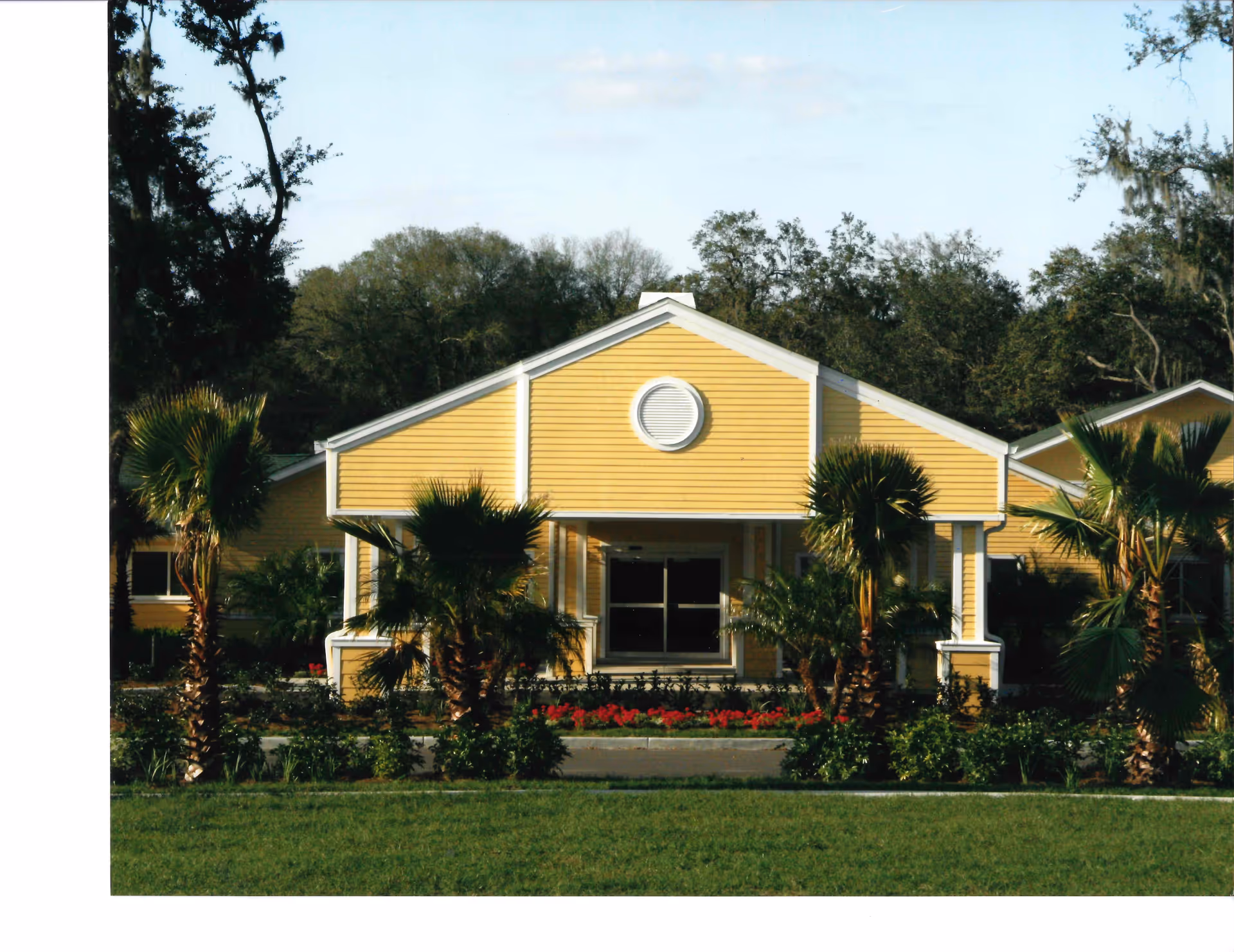 Front exterior view of a single-story yellow building with white trim, surrounded by palm trees and landscaping, under a clear sky.