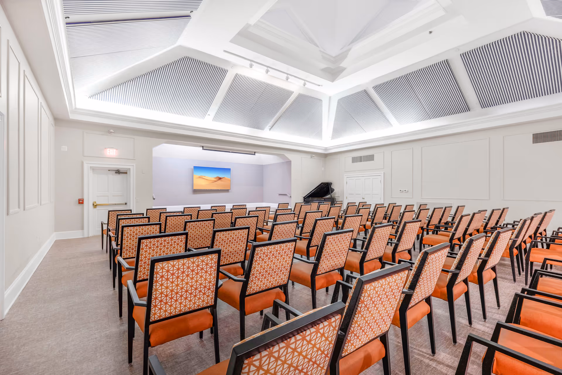 A bright, spacious meeting or presentation room with rows of orange cushioned chairs featuring patterned backs, facing a wall-mounted screen displaying a desert scene. The room has a high ceiling with recessed lighting and a grand piano in the corner.