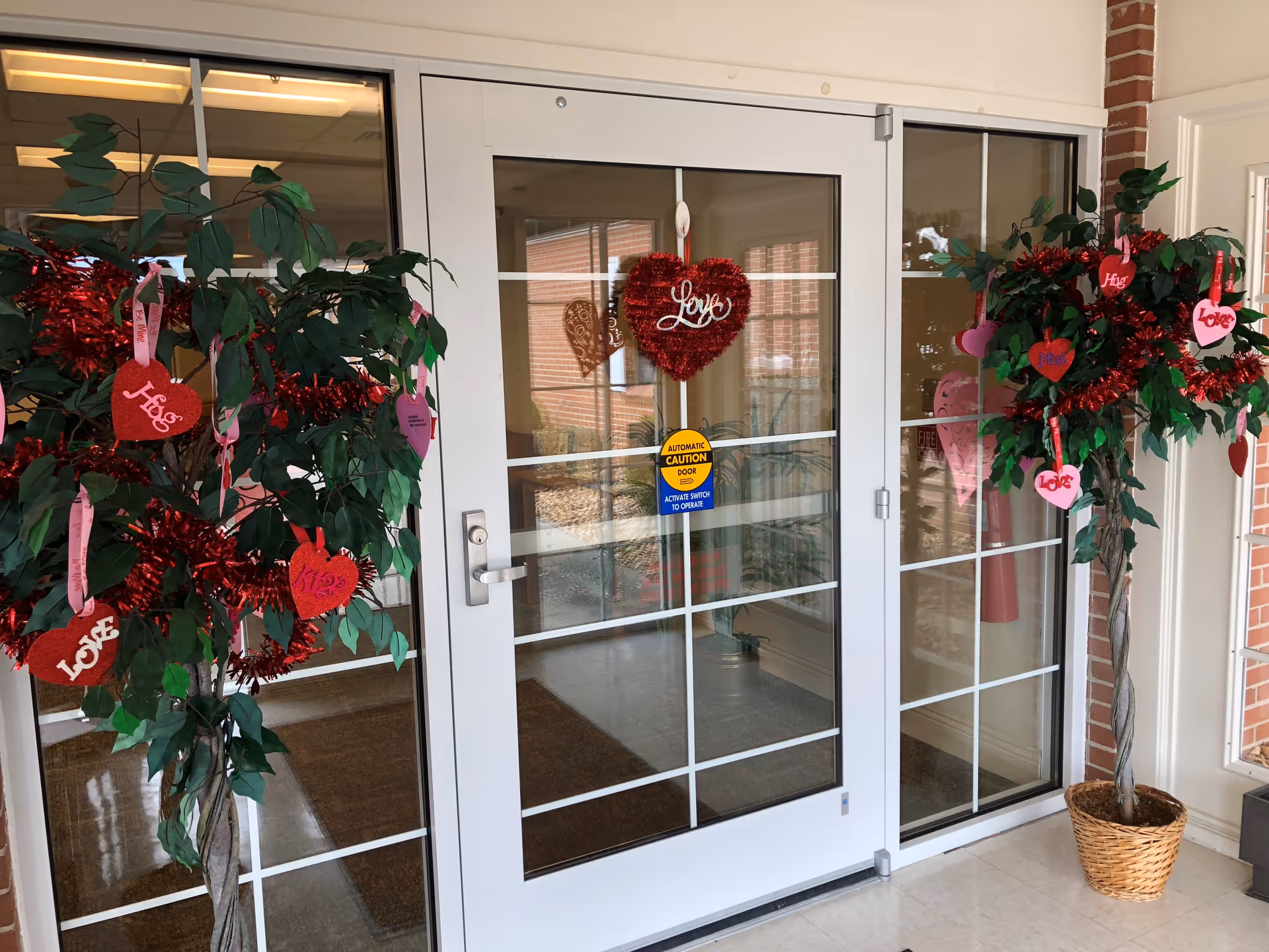 Glass automatic double doors decorated with red and pink Valentine's Day heart ornaments and garlands, flanked by two potted artificial trees also adorned with heart decorations. The doors have a caution sticker indicating they are automatic and require activation to open.